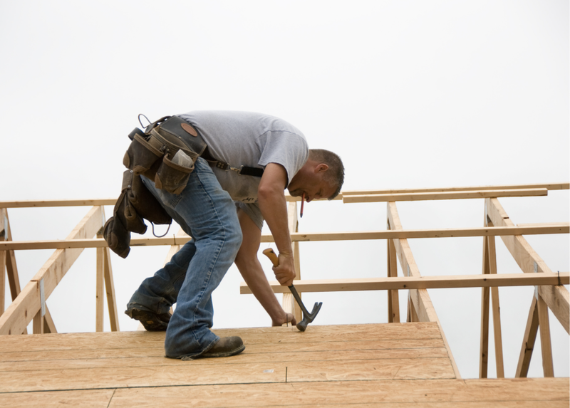 Construction worker hammering wood on a roof, wearing work boots, and a tool belt.