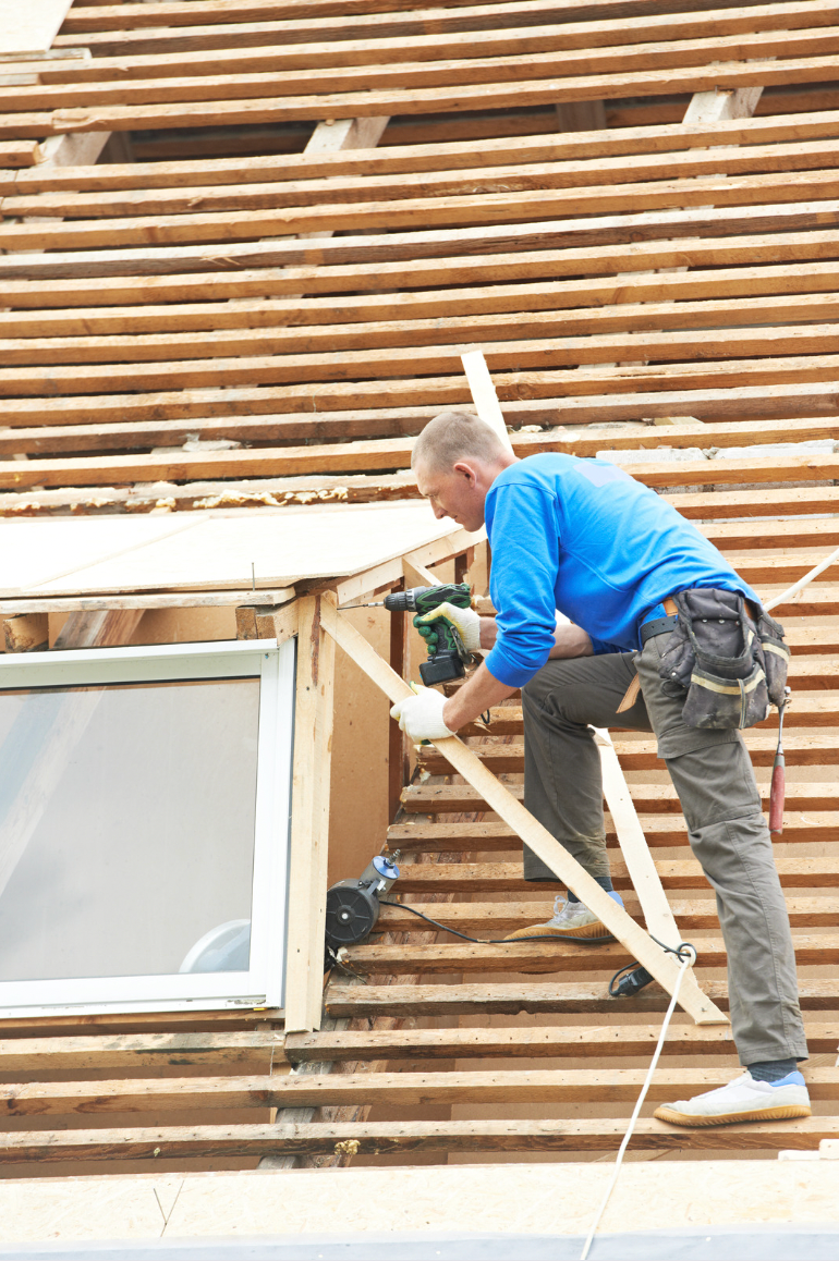 Construction worker in blue shirt, installing wood trim around a skylight on a partially constructed roof.