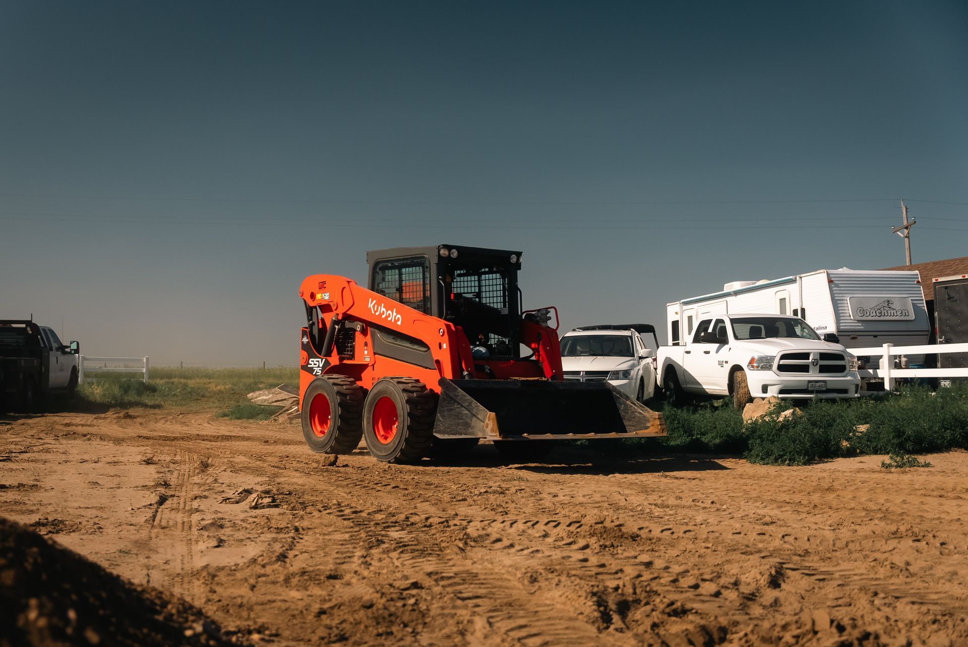 Orange skid steer loader on a dirt lot, parked near white vehicles.