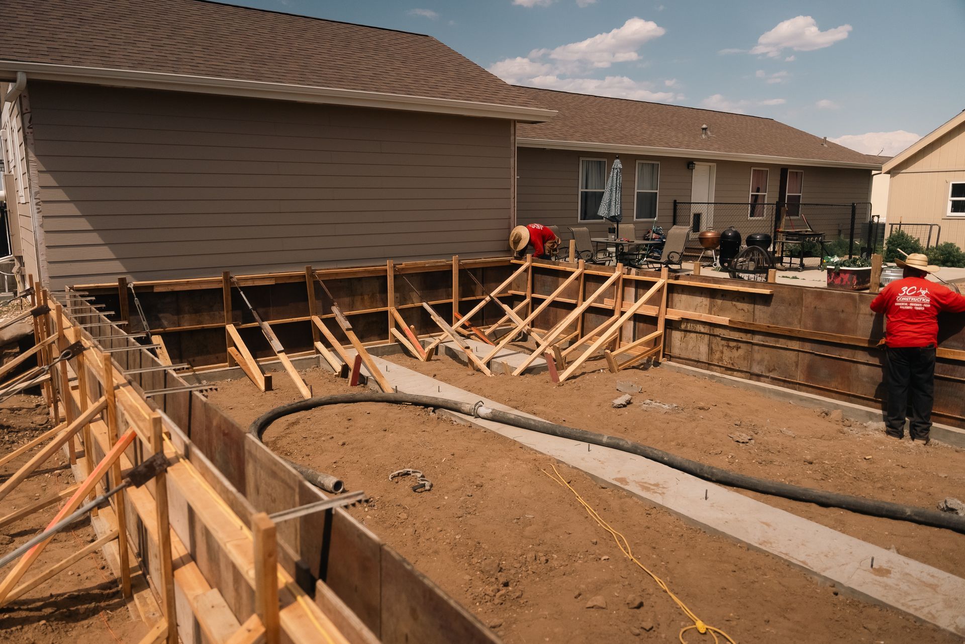 Construction site with wooden forms for concrete foundation near a house; worker in red jacket.