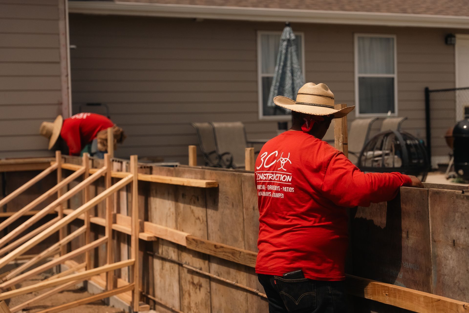 Two construction workers in red shirts and cowboy hats building a wooden structure outdoors.