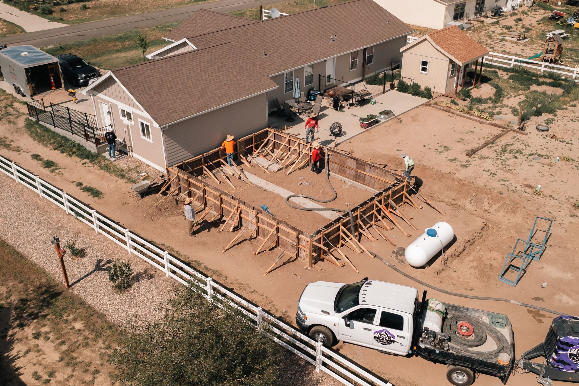 Construction site with workers, forms, and concrete pouring. A white truck and propane tank are visible.