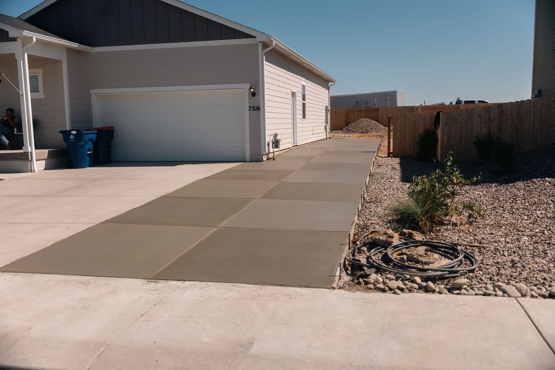 Driveway of a house with recently poured concrete squares. Blue trash bins by the garage. Sunny day.
