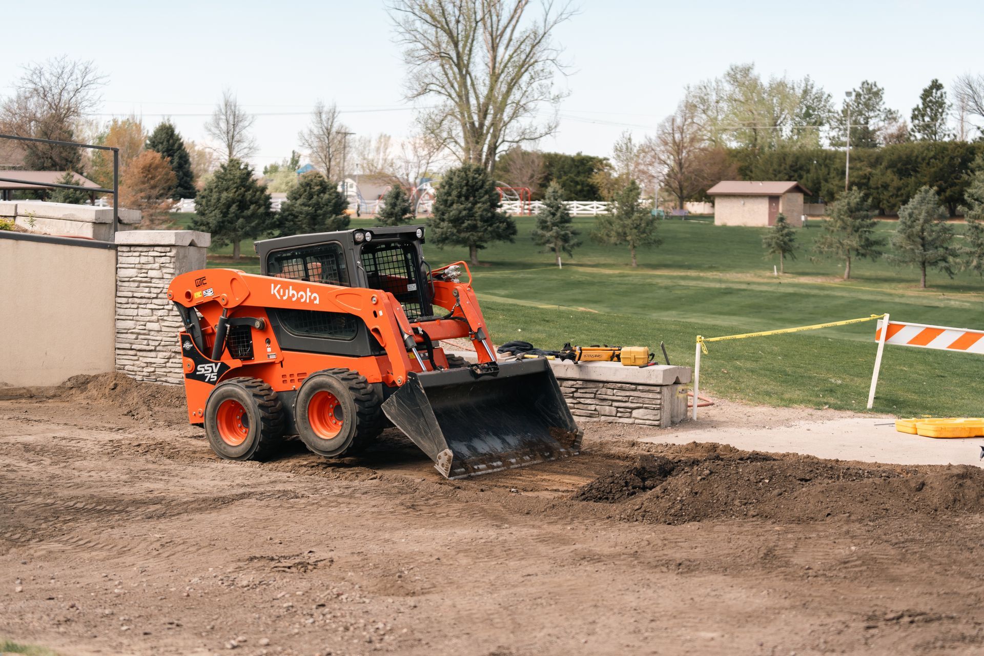 Orange Kubota skid steer digging in dirt near a wall and lawn.