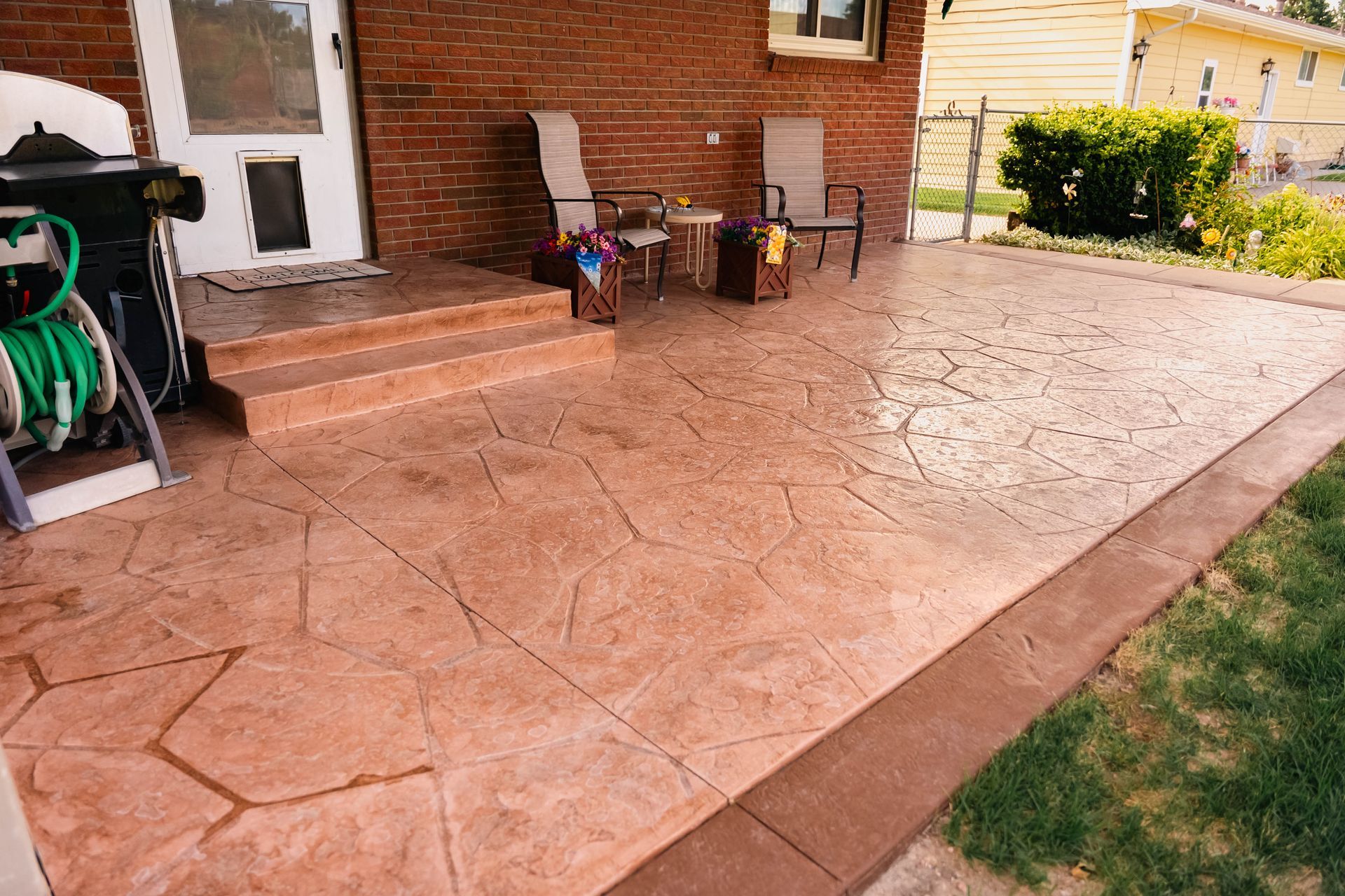 Stamped concrete patio, reddish-brown color, next to brick house with chairs and greenery.