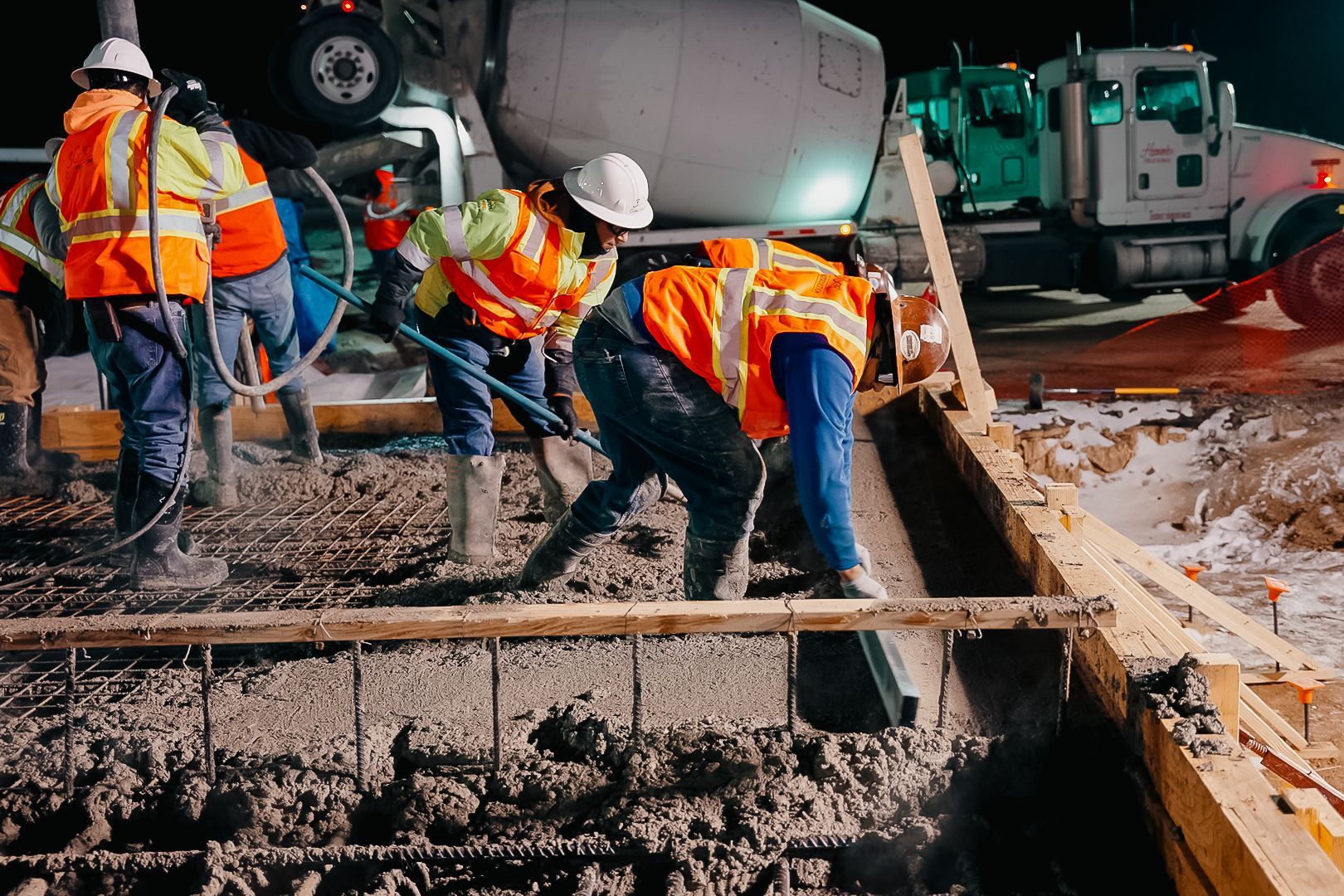 Construction workers pouring concrete at night. Workers wear safety vests and helmets, near a concrete truck.
