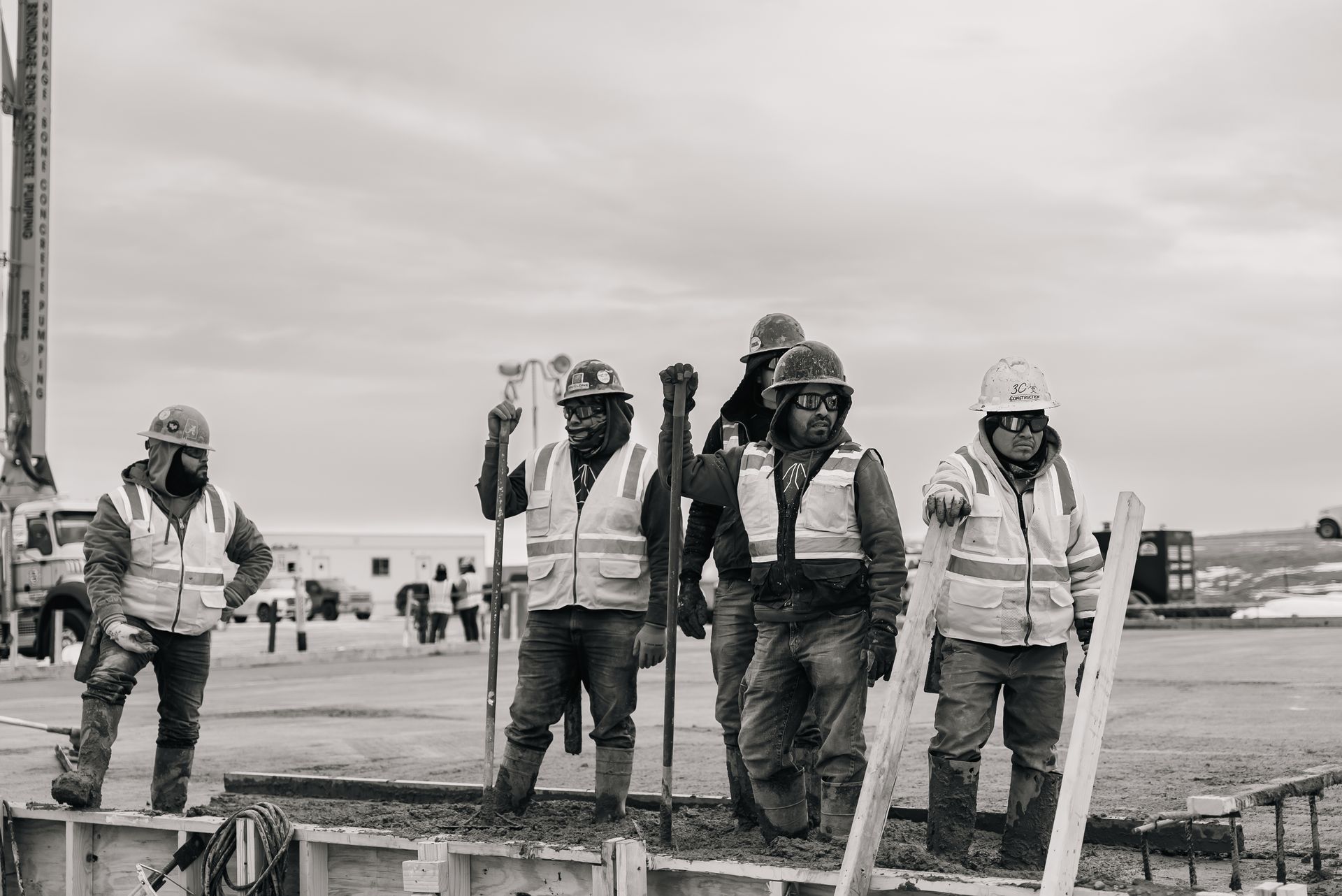 Construction workers in safety vests and hard hats stand on a concrete form, cloudy sky.