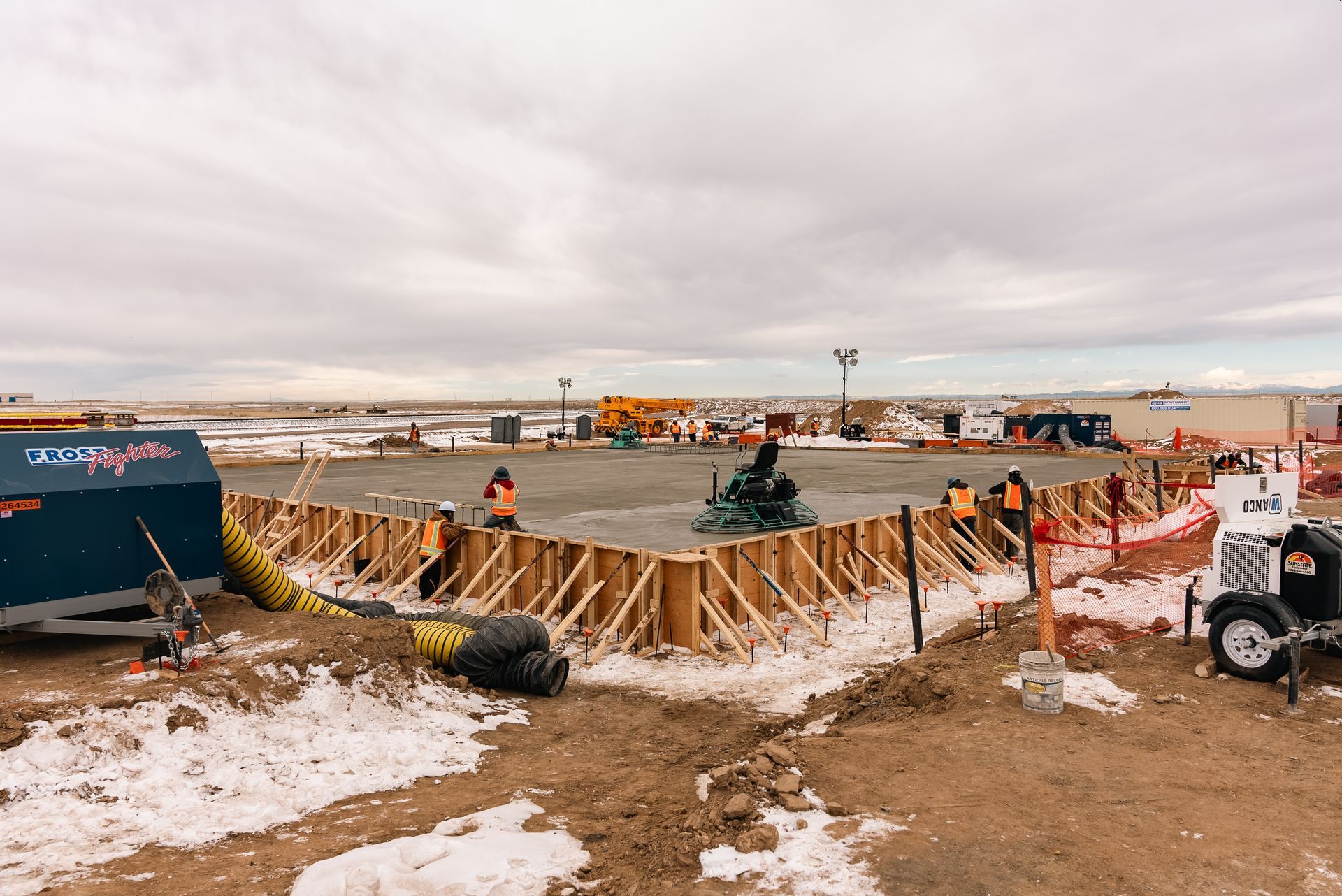 Construction site, concrete being poured into wooden forms, workers present, cloudy sky.