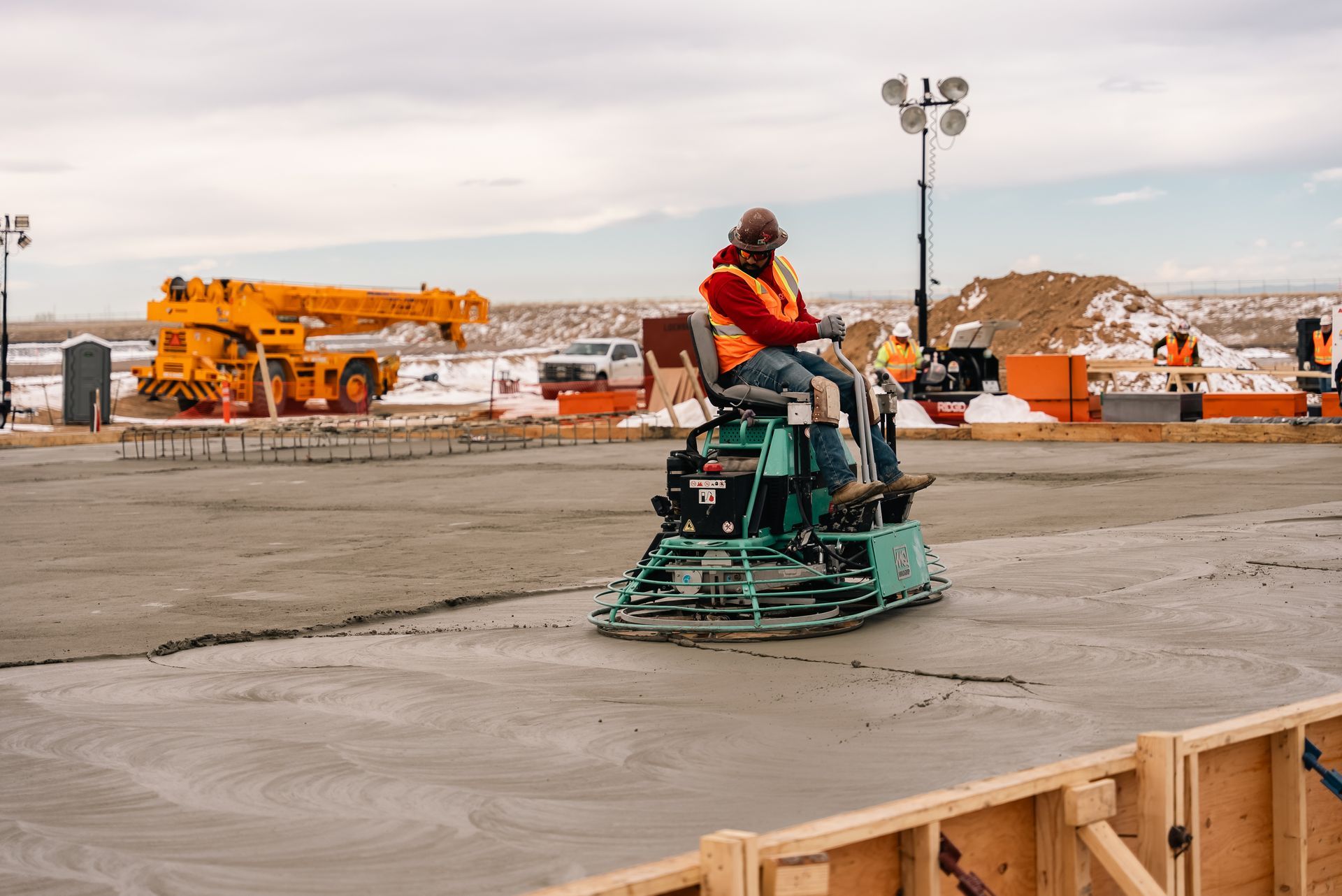 Person operating a ride-on power trowel on wet concrete at a construction site. Orange safety vest, outdoor setting.
