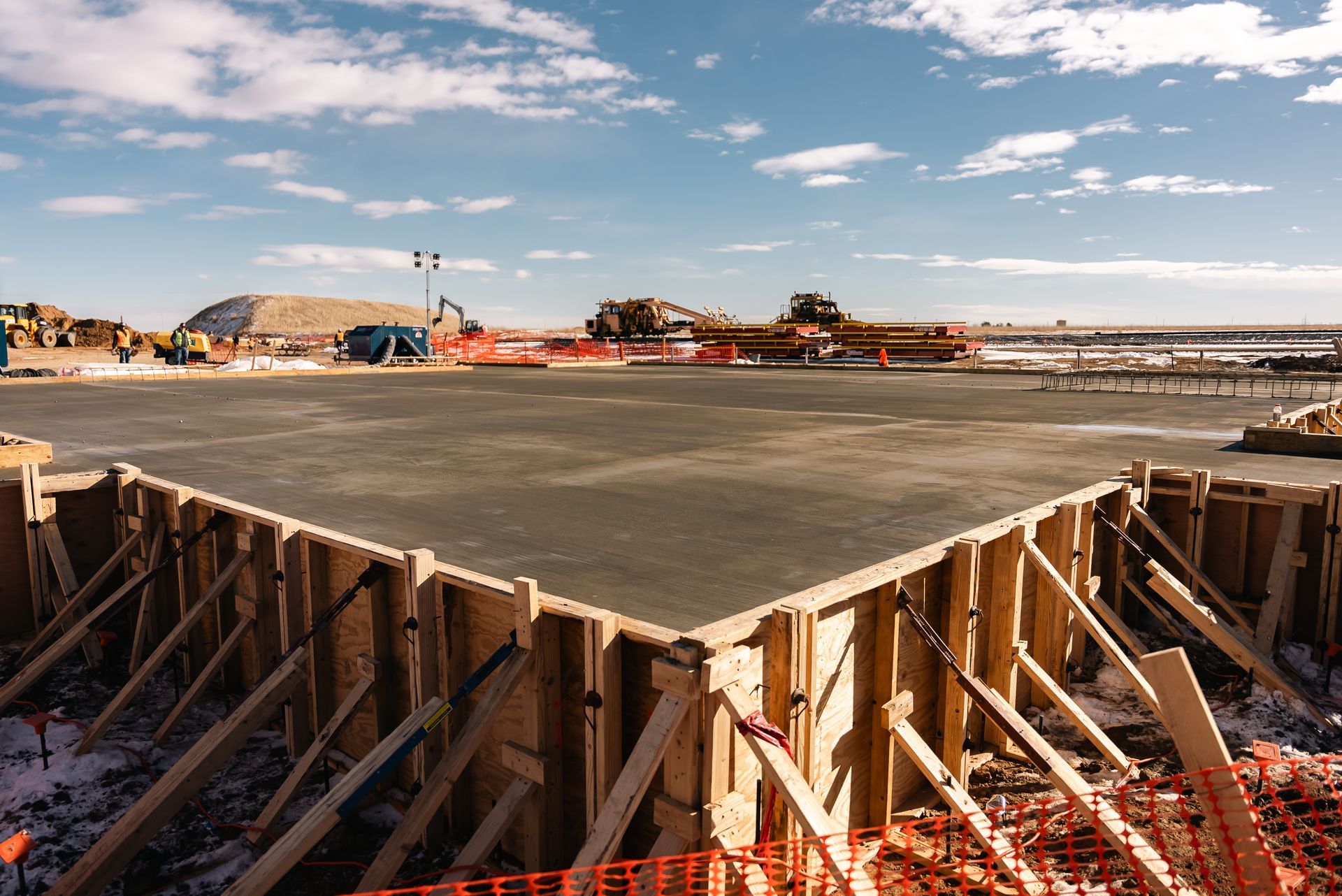 Concrete foundation being poured, with wooden forms in foreground and construction site in background, under blue sky.