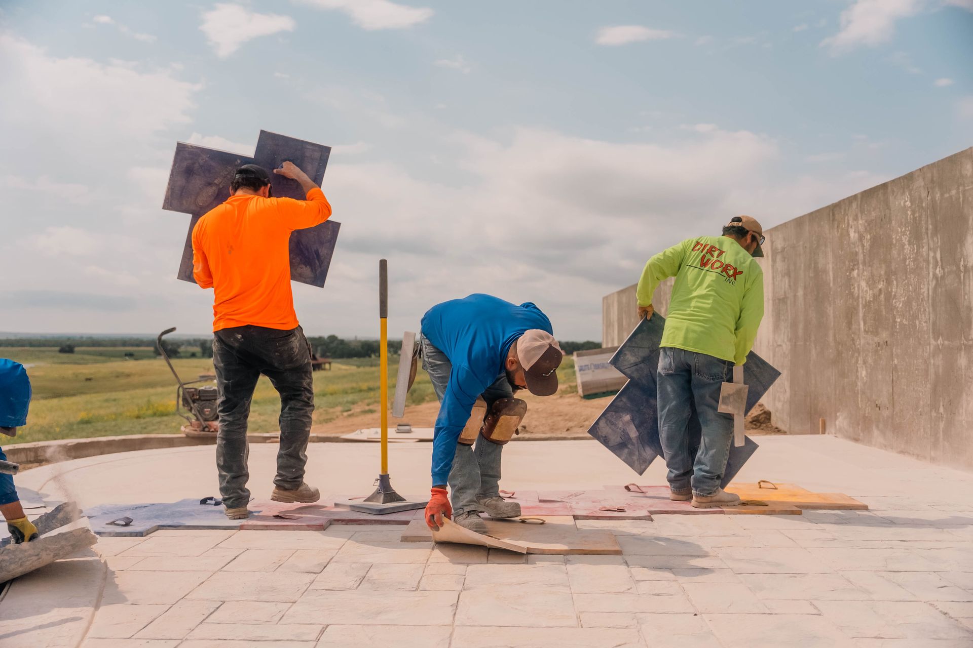 Construction workers laying patterned tiles on a concrete surface outside, bright sky above.