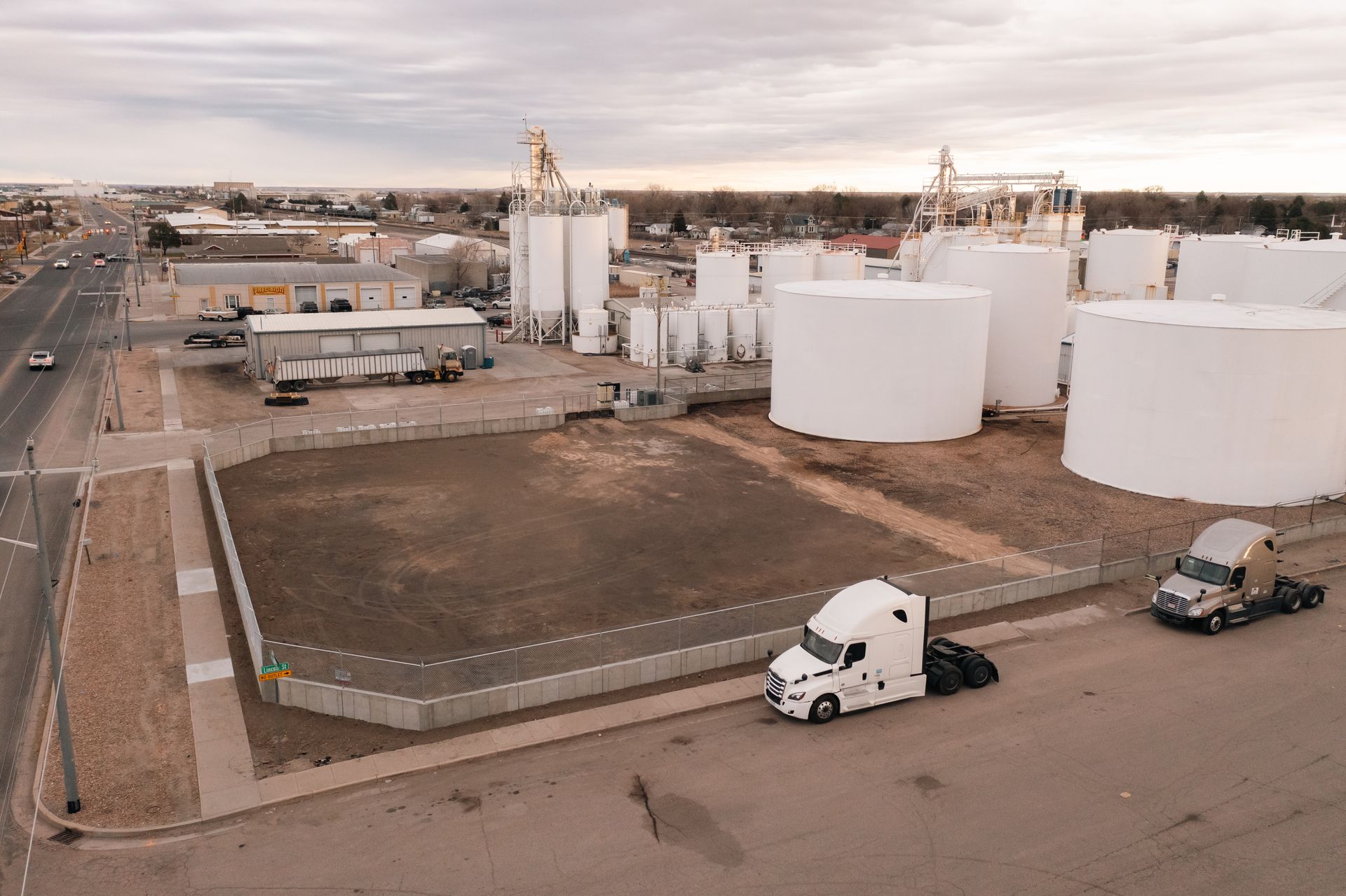 Aerial view of an industrial facility with large white storage tanks and two semi-trucks on a roadway.