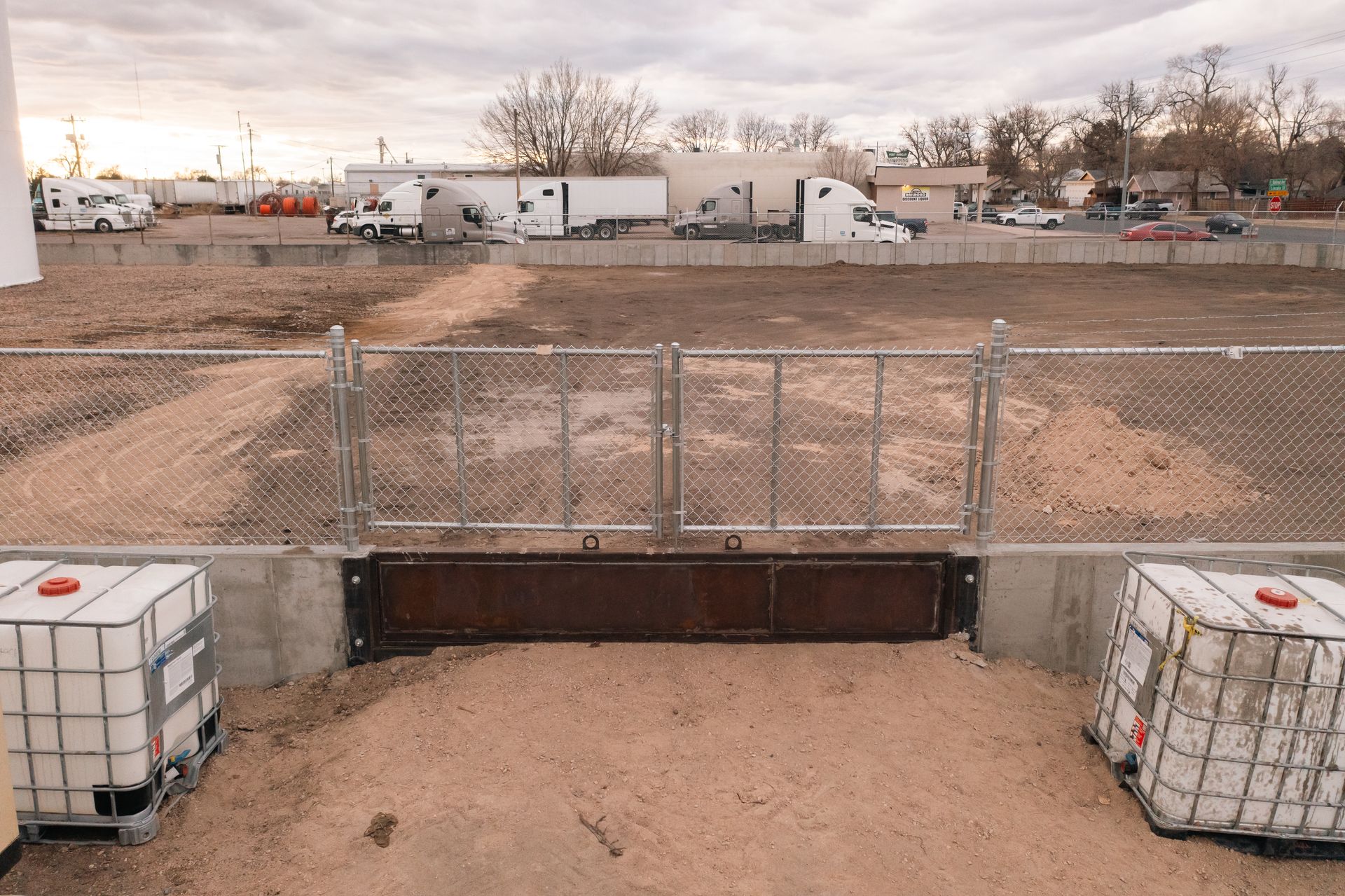 Chain link fence gate with metal barrier, dirt area, and storage containers. Industrial setting with trucks visible.