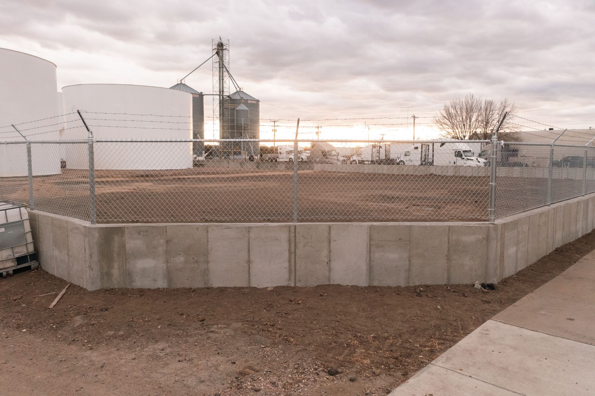 Industrial facility with concrete retaining wall, chain link fence, and silos under cloudy sky.