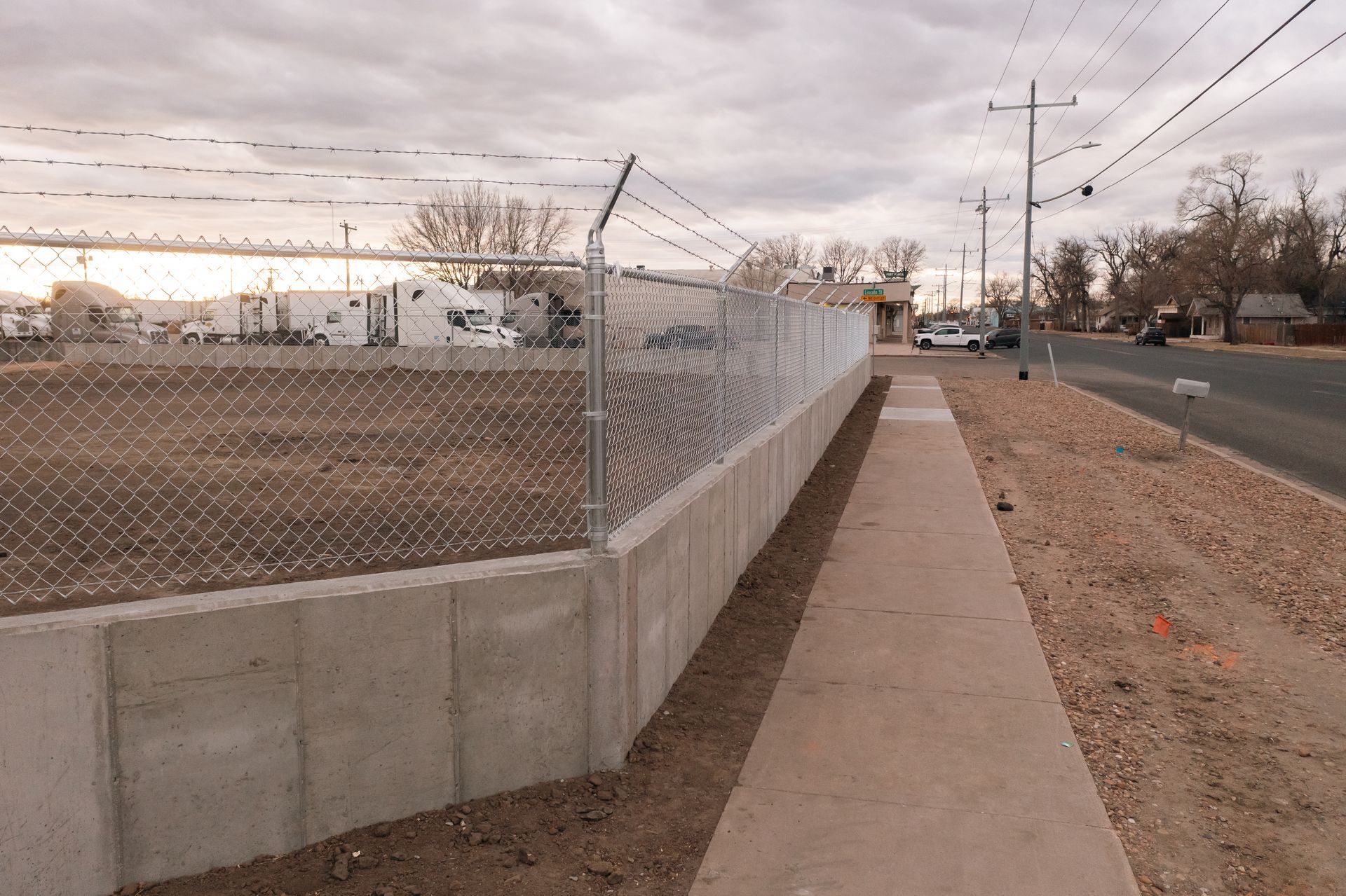 Concrete retaining wall topped with chain-link fence and barbed wire next to a sidewalk and road.
