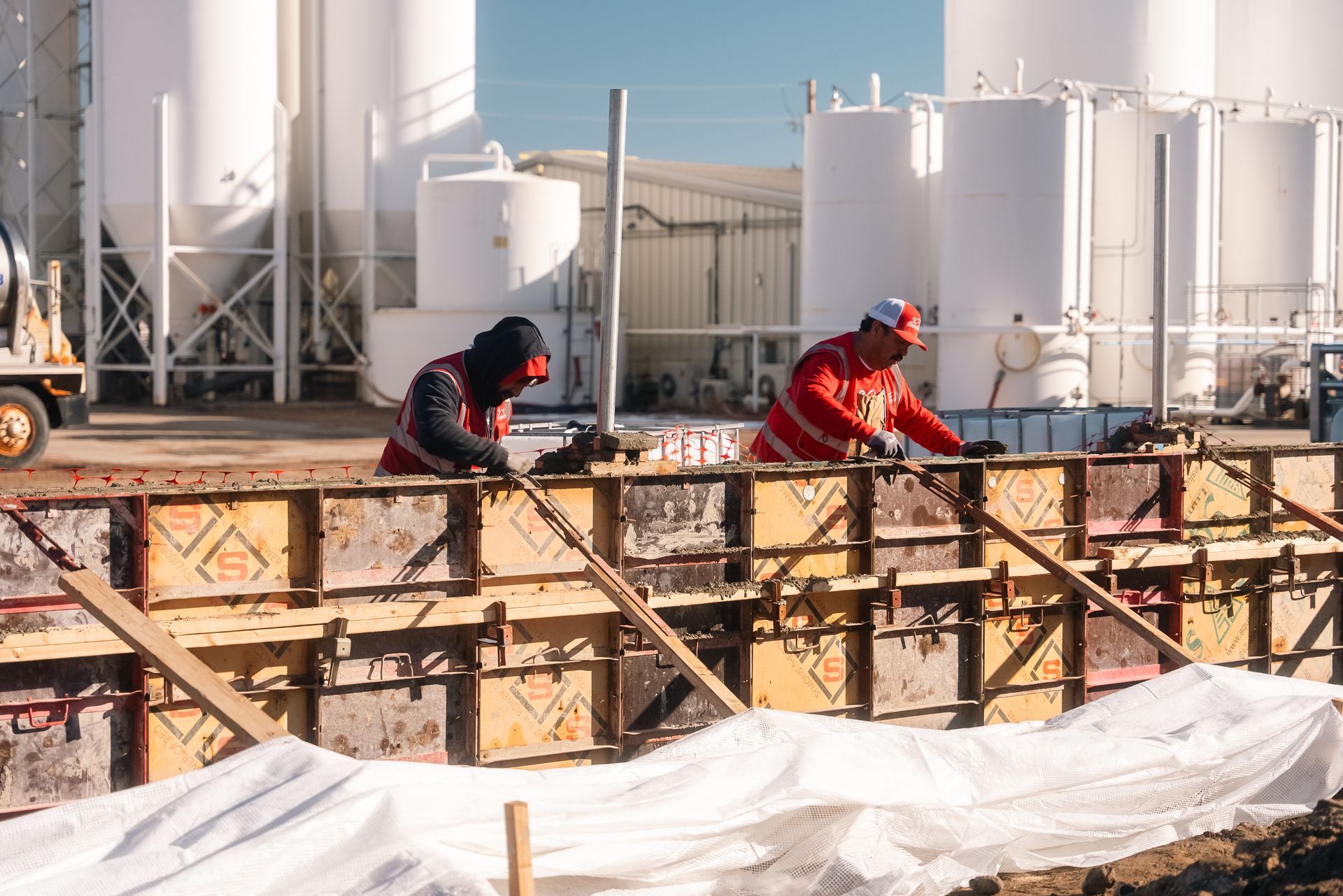 Two construction workers in red vests building a concrete form outside industrial tanks.