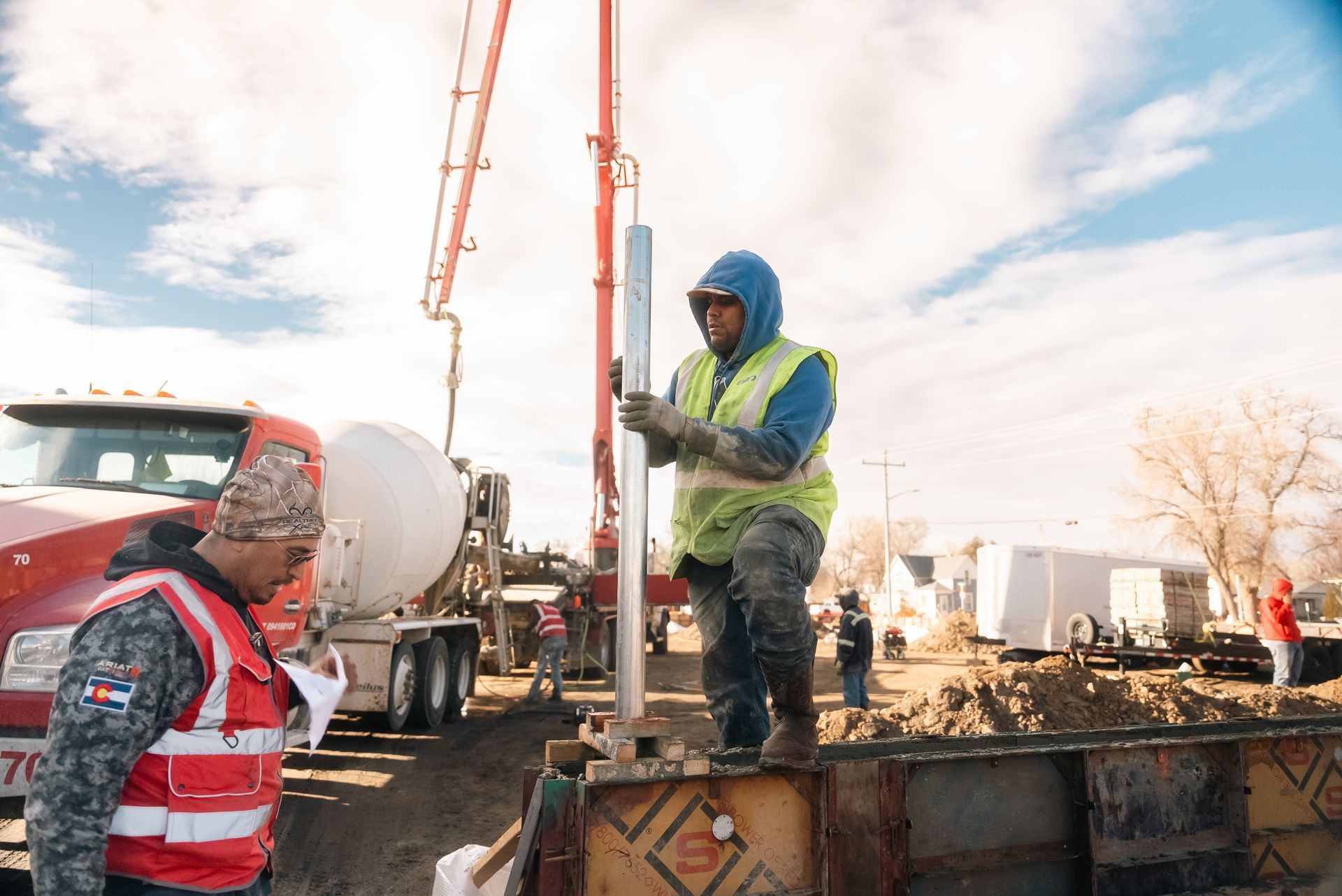 Construction workers at a concrete pour: one in a vest holds a metal rod, the other looks on. 