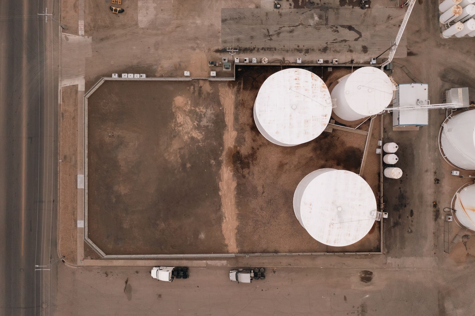 Overhead view of an industrial site with large white tanks and a rectangular dirt area, near a road and trucks.