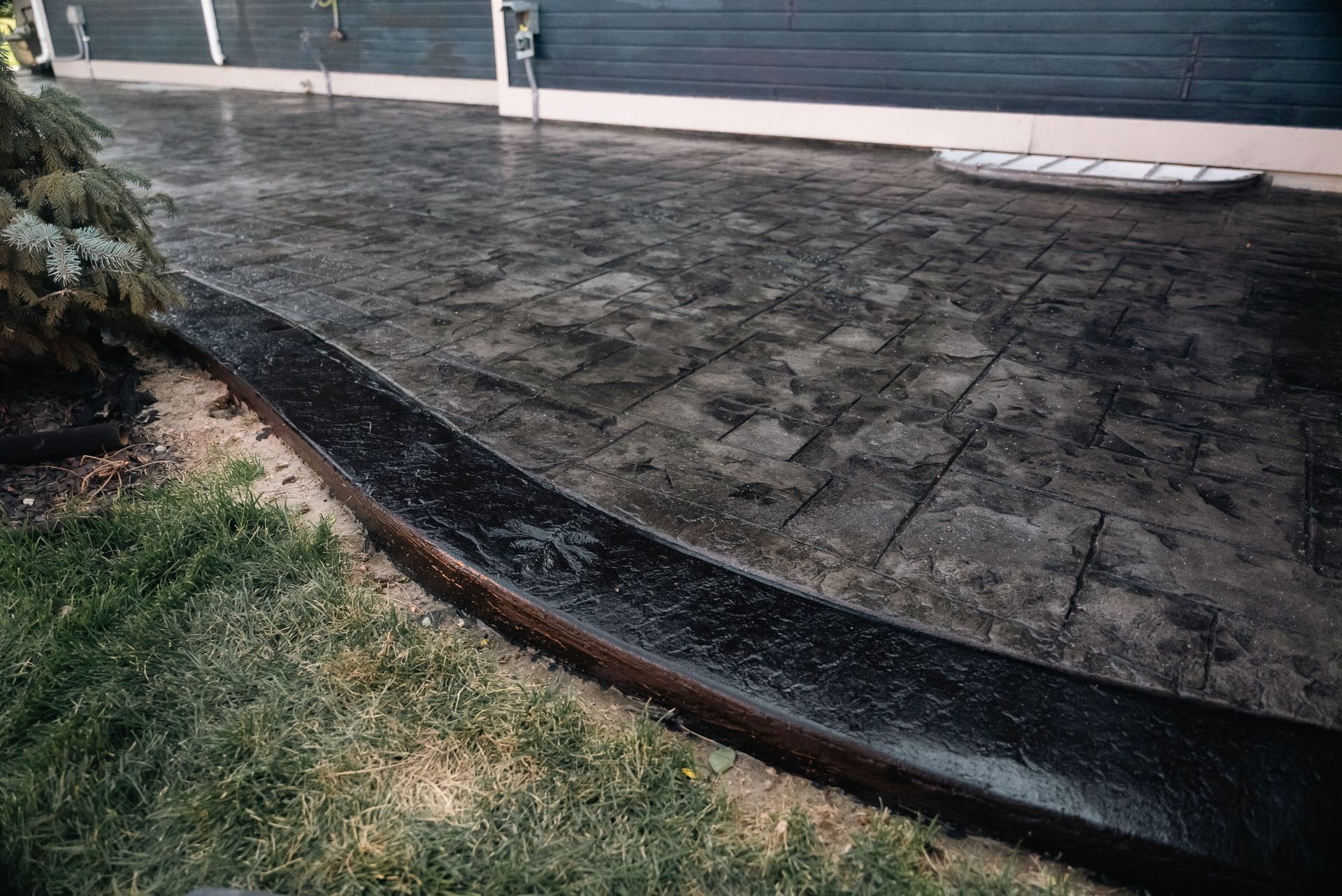 Dark, wet stamped concrete patio with curved border, beside grass. Building and window well in background.