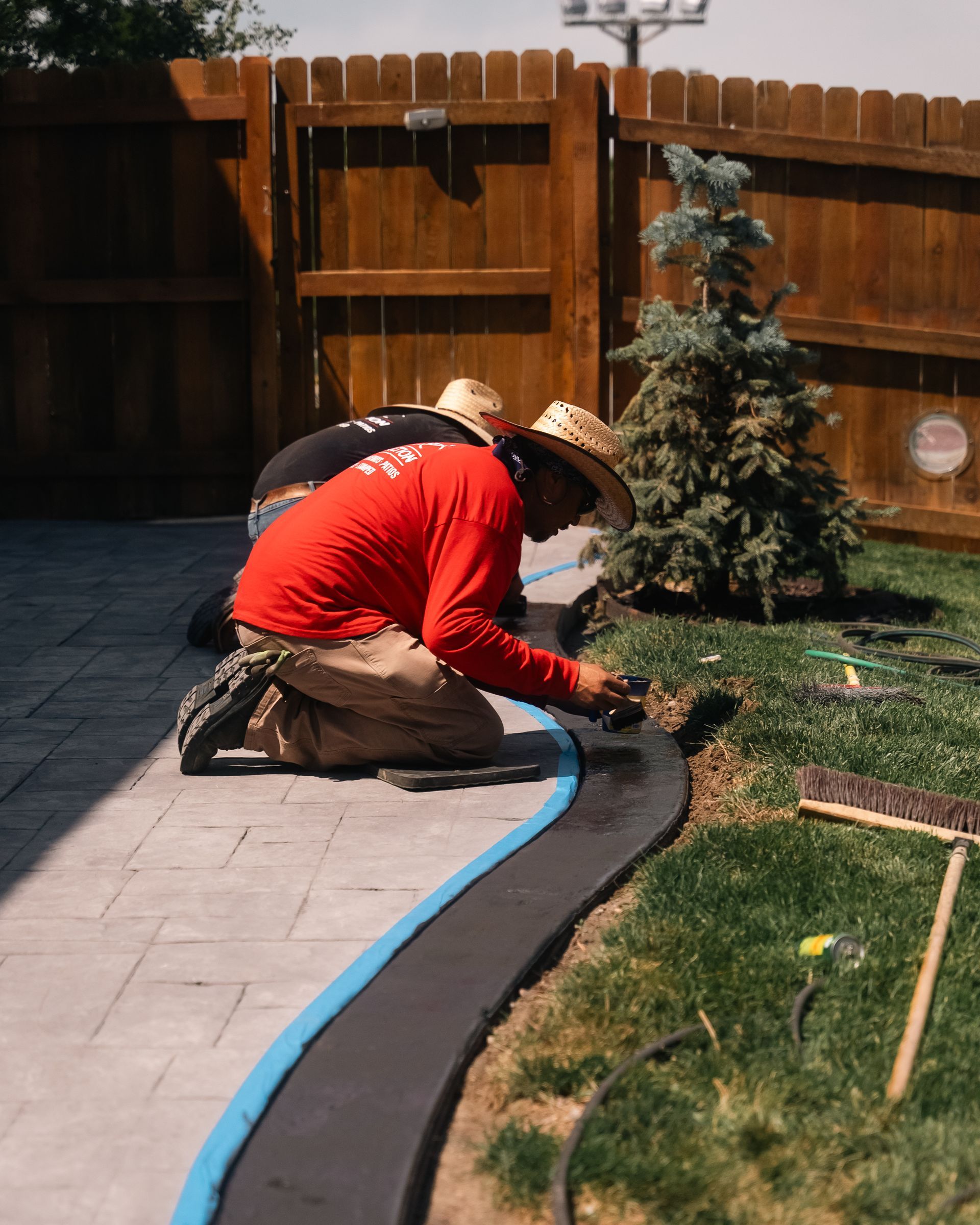 Two workers install a dark border along a curvy walkway. One kneels, applying material, near a lawn.