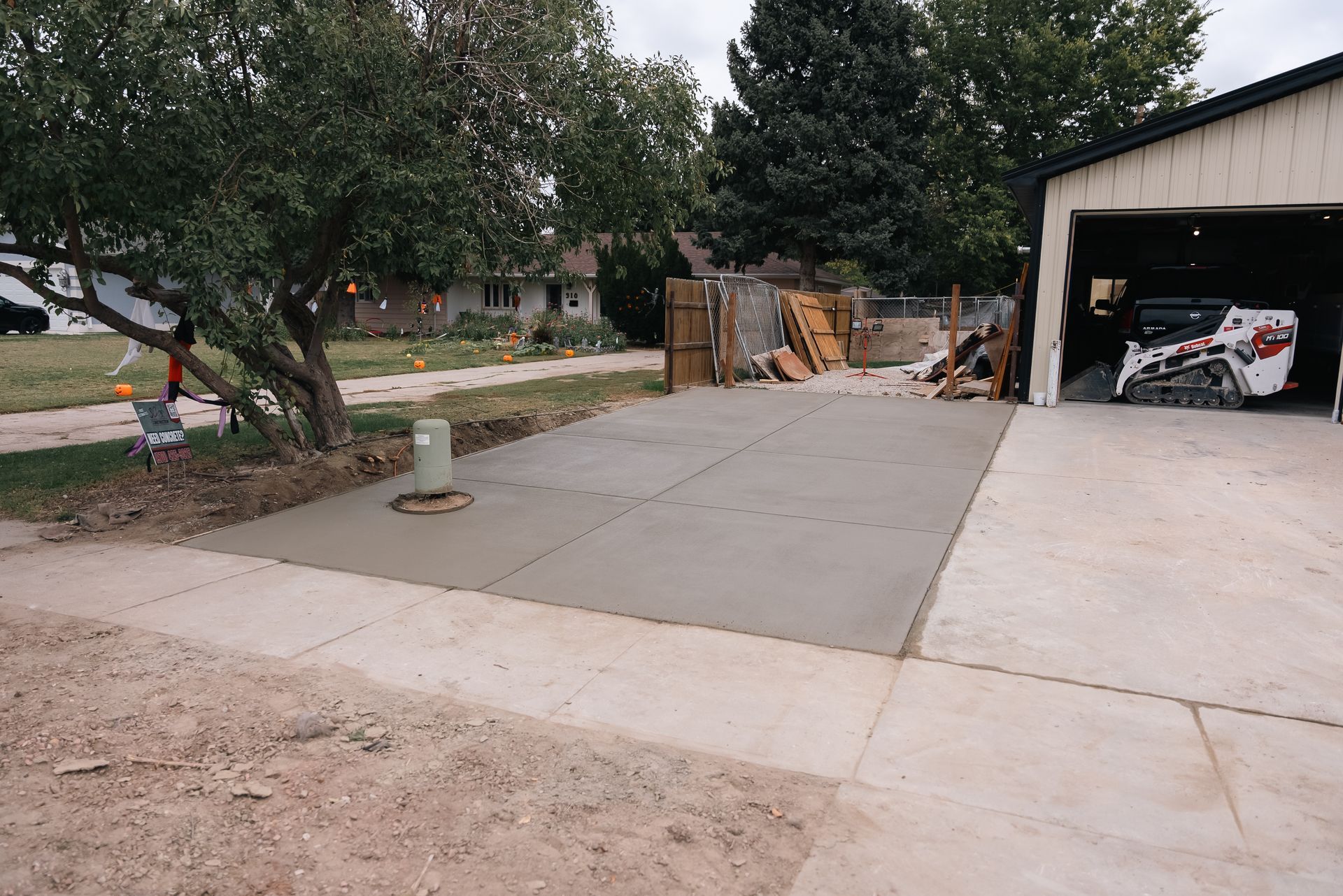 Freshly poured concrete driveway adjacent to a garage and house. A small tree and construction materials are visible.