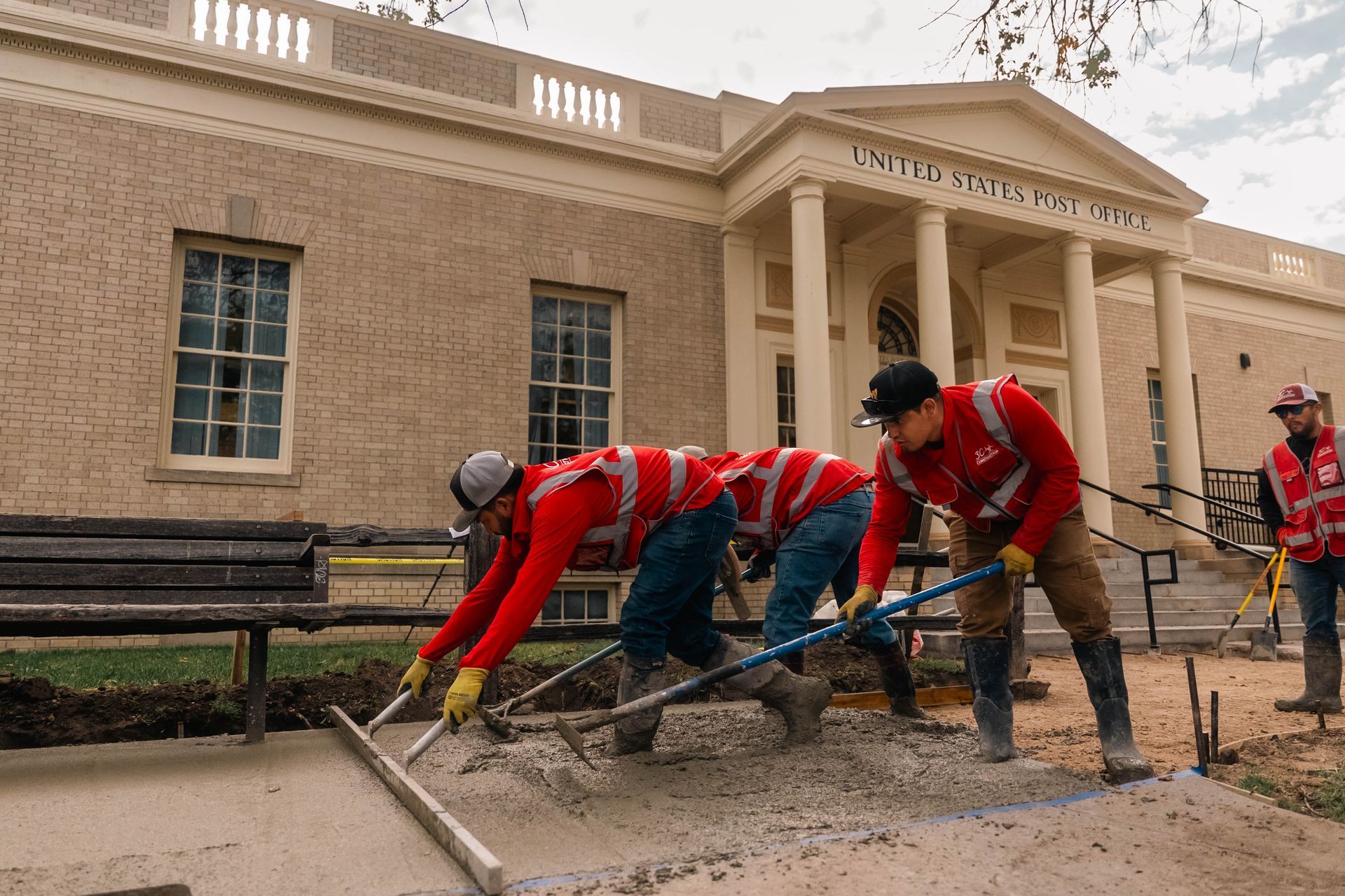Construction workers leveling wet concrete in front of a building with columns; cloudy day.