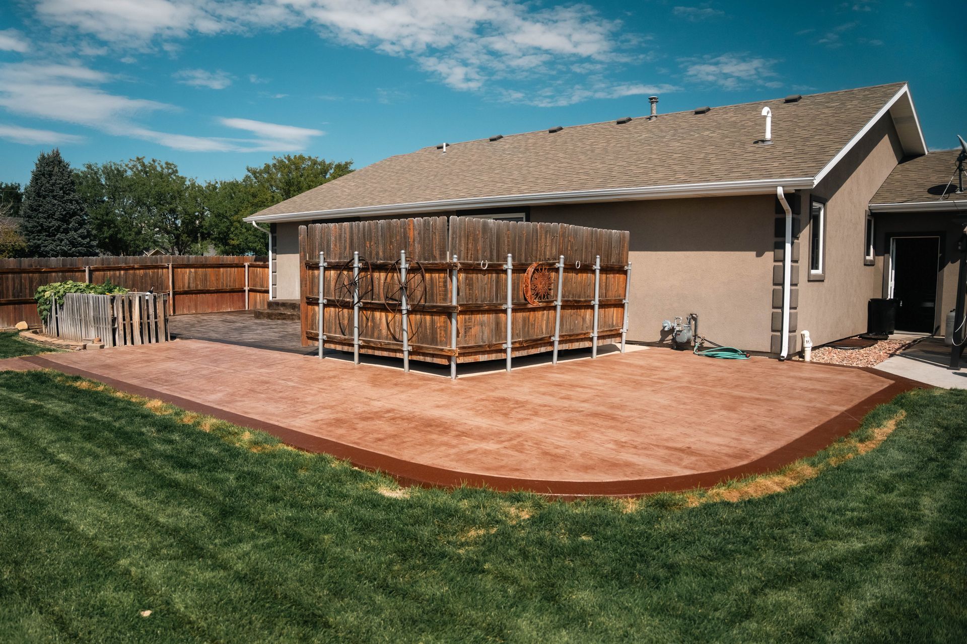 Backyard patio with brown concrete, wooden fence, house, and green lawn.