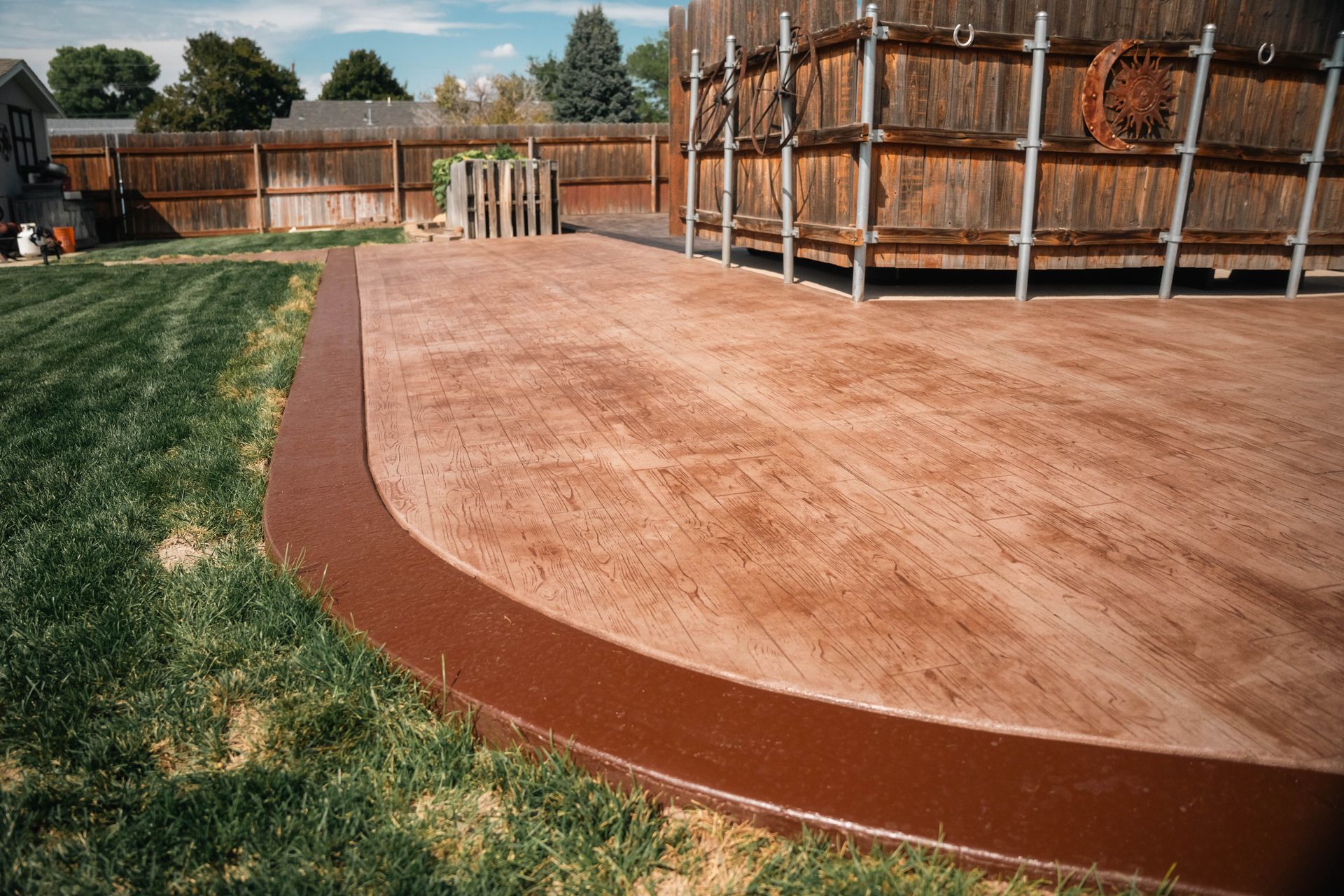 Brown concrete patio with a dark brown border next to a lawn and wooden structure.