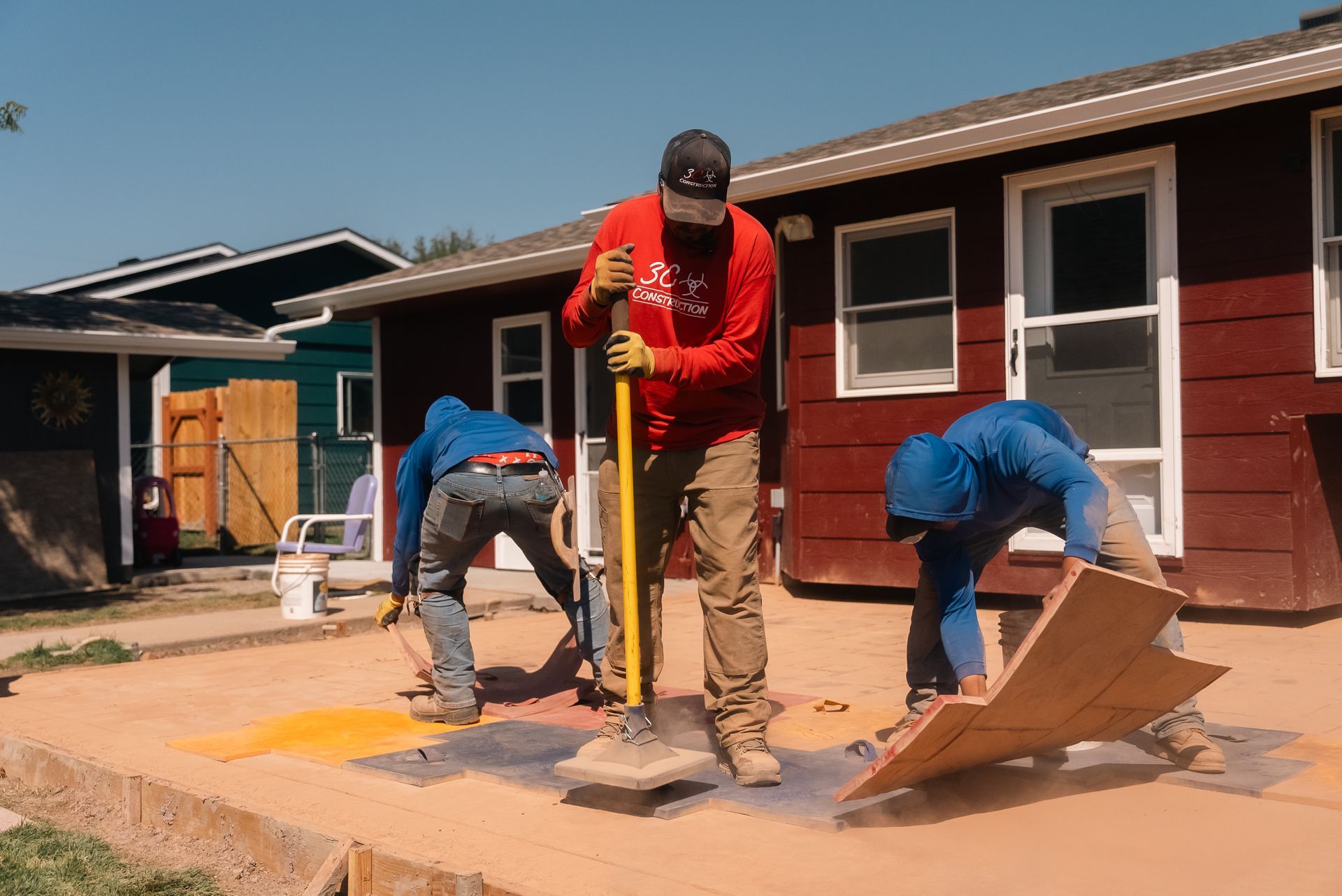 Three workers on a sunny day repairing a concrete patio; one uses a shovel, others work on the surface near houses.