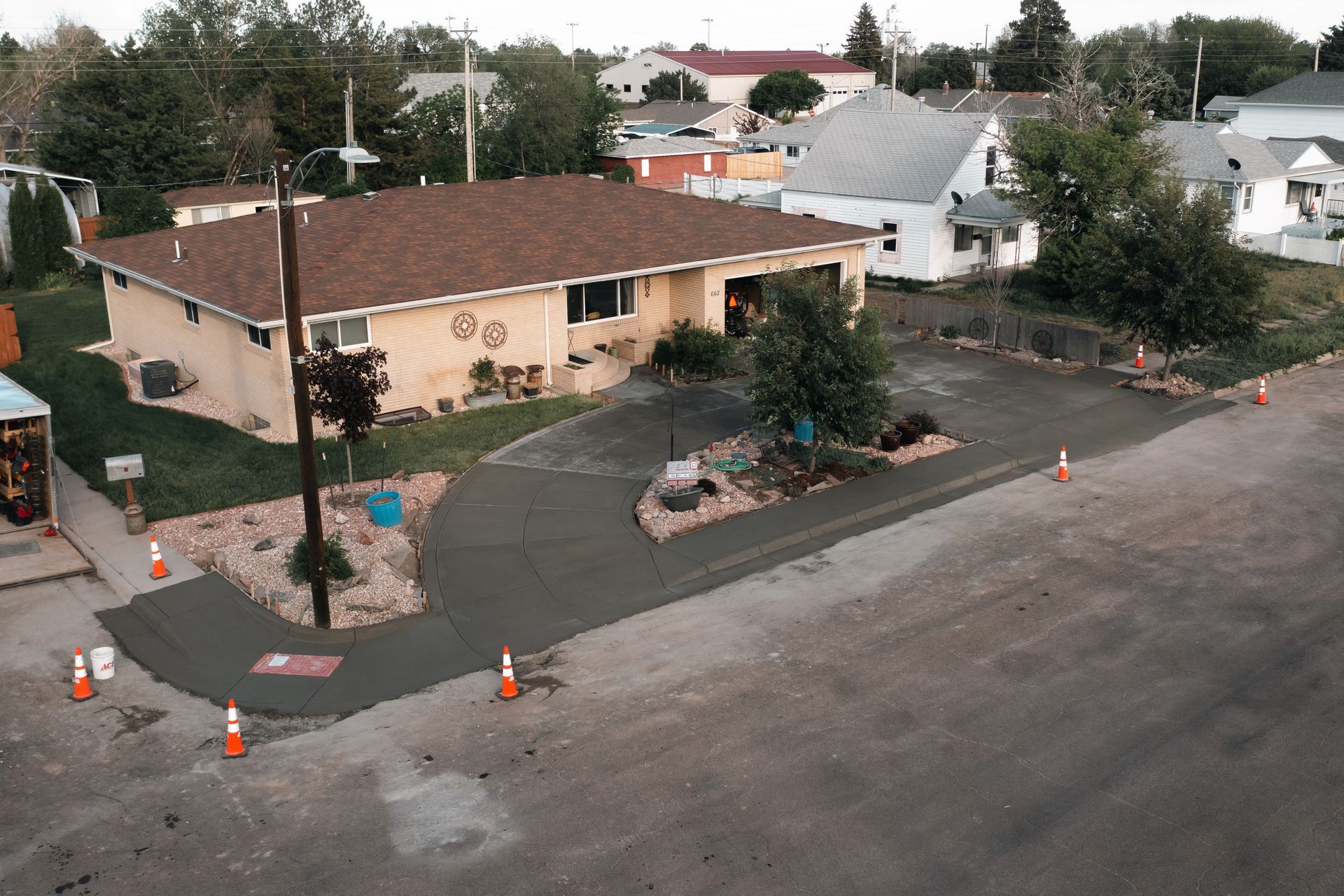 Asphalt driveway in front of a tan house. Cones mark the perimeter. Green lawn and a few trees.