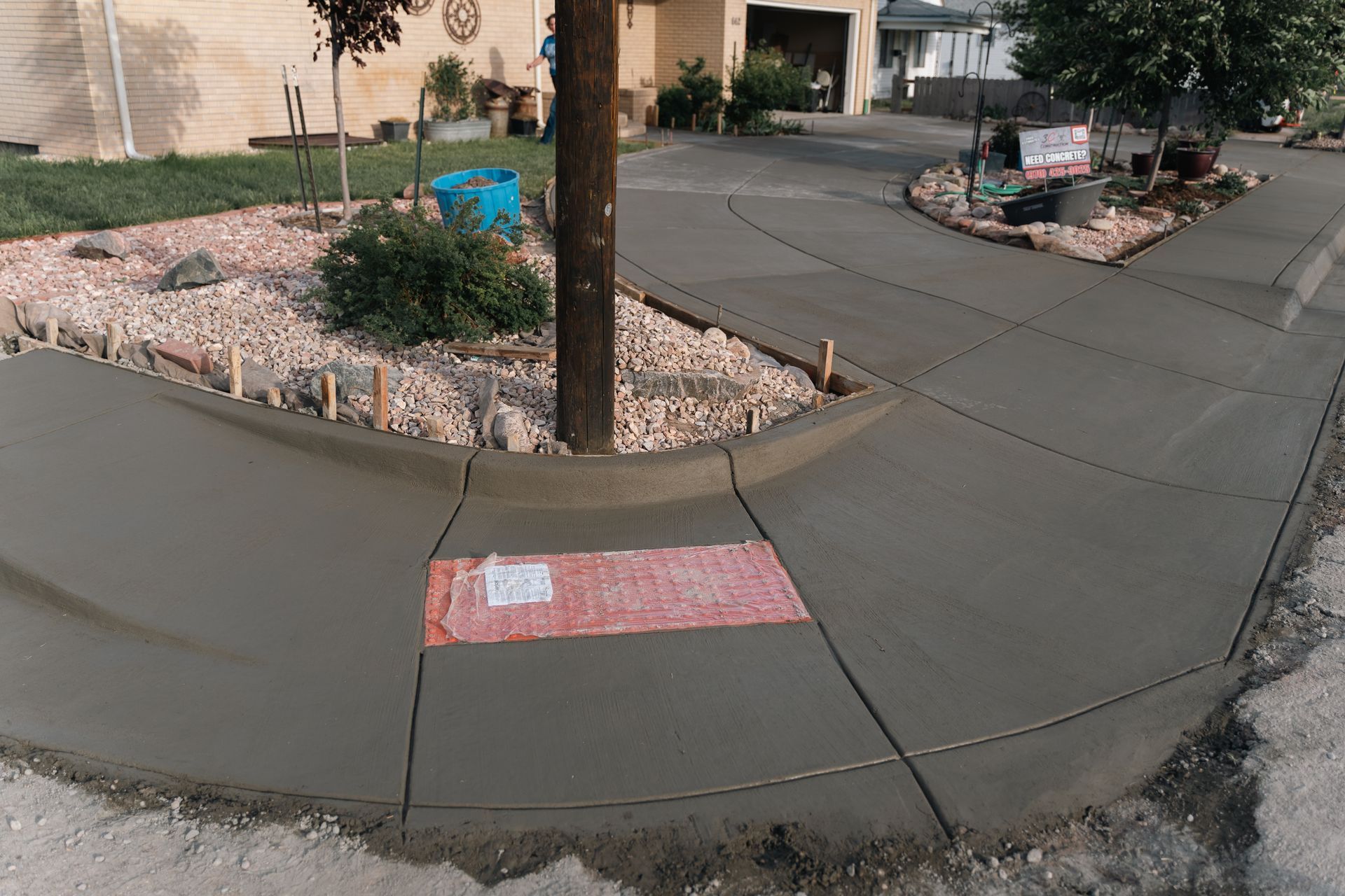 Freshly poured concrete sidewalk and curb around a tree, with a red mat and building in the background.