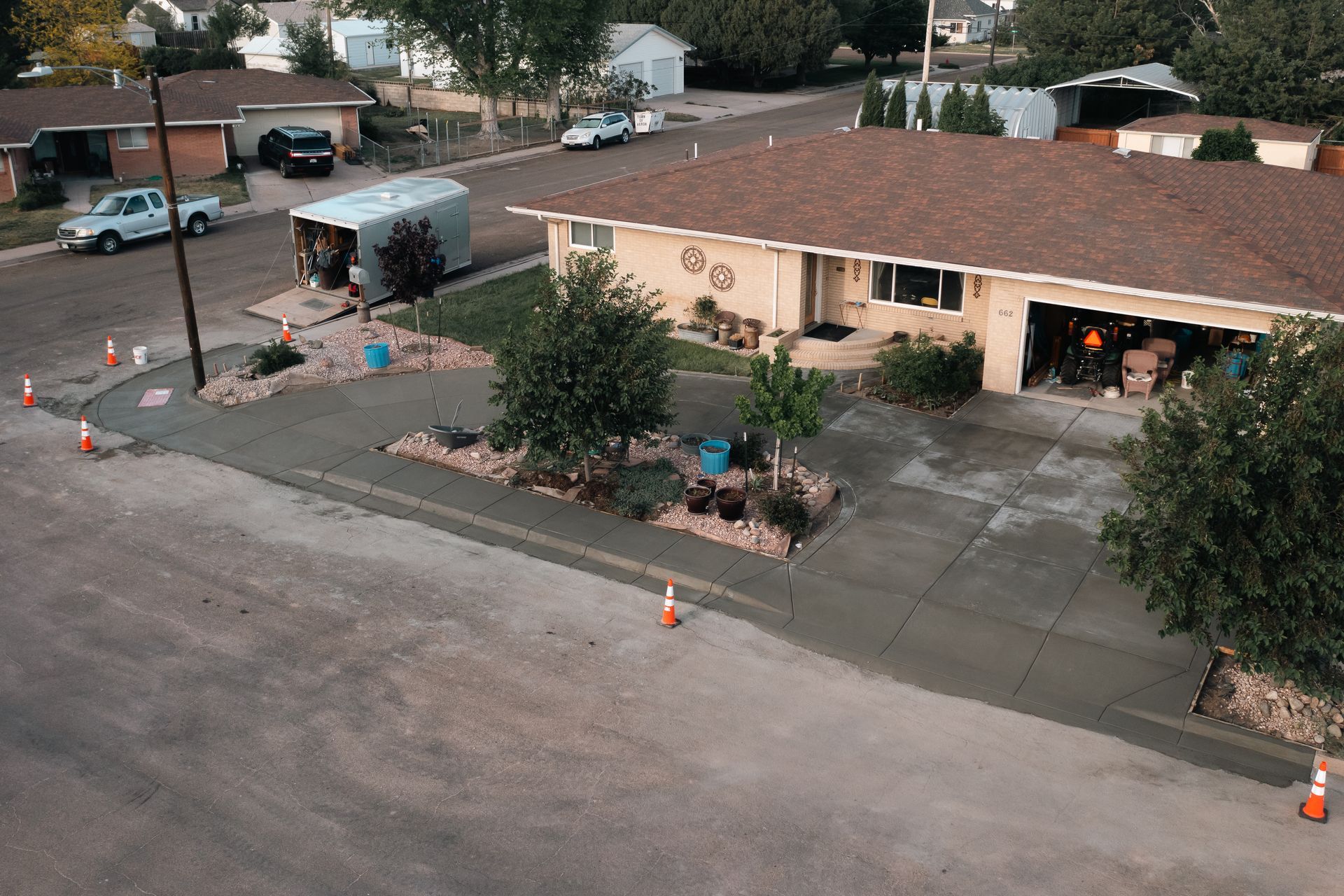 Residential street scene; beige house with brown roof, driveway, and landscaping; construction cones visible.