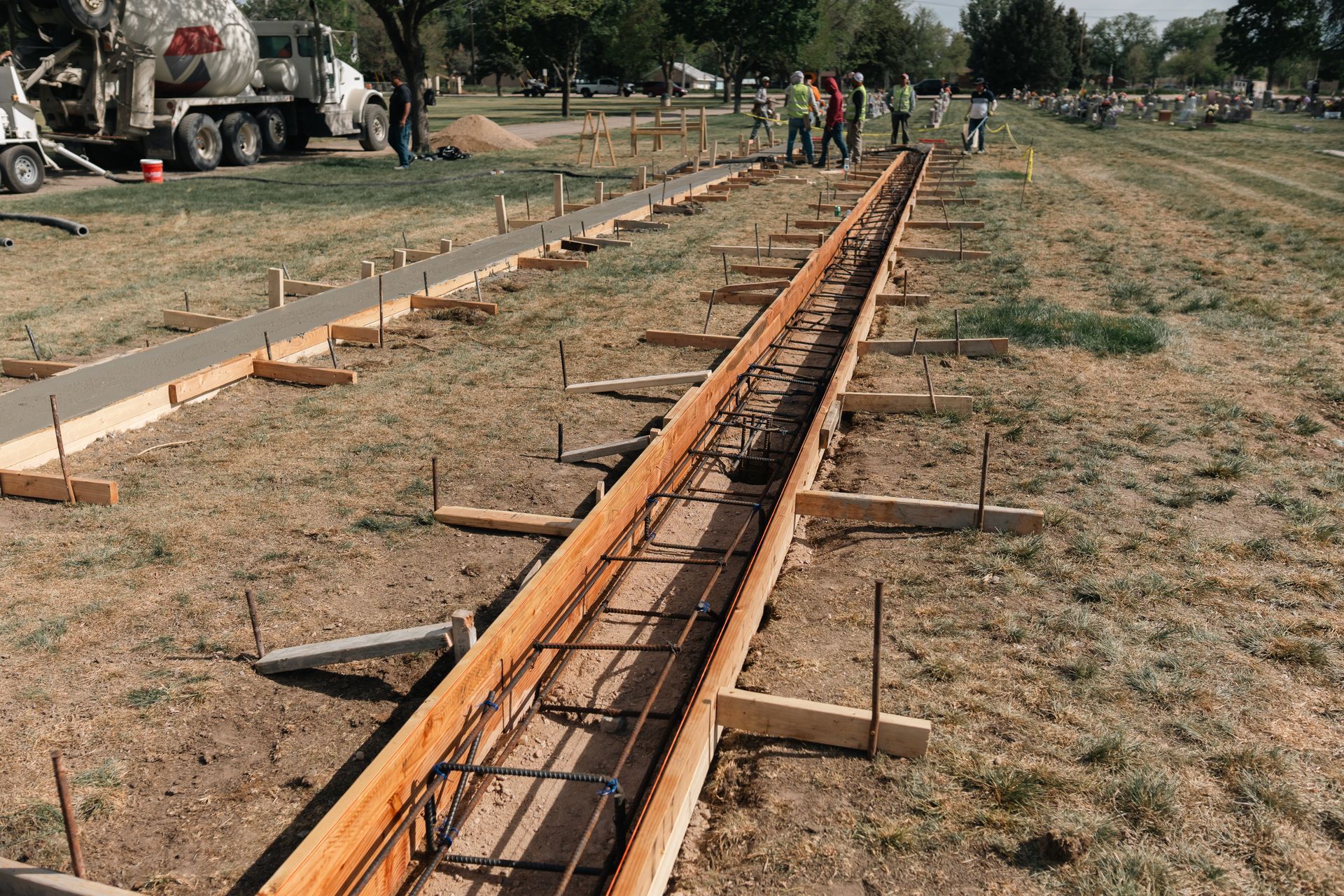 Construction site: wooden forms and rebar prepared for concrete. Cement truck and workers in the background.