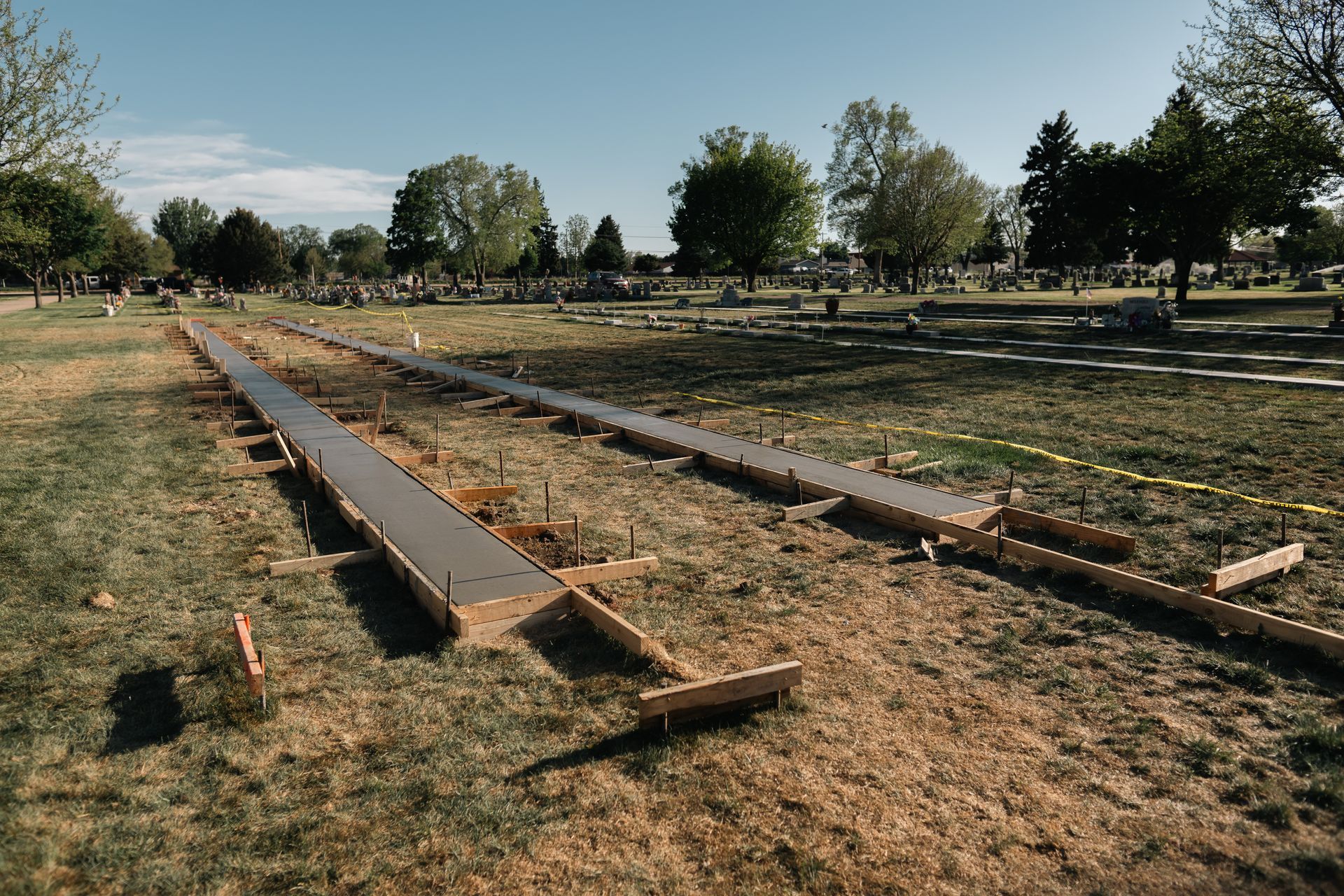 Two concrete forms are set in a grassy field, possibly for a walkway, with a cemetery in the background.