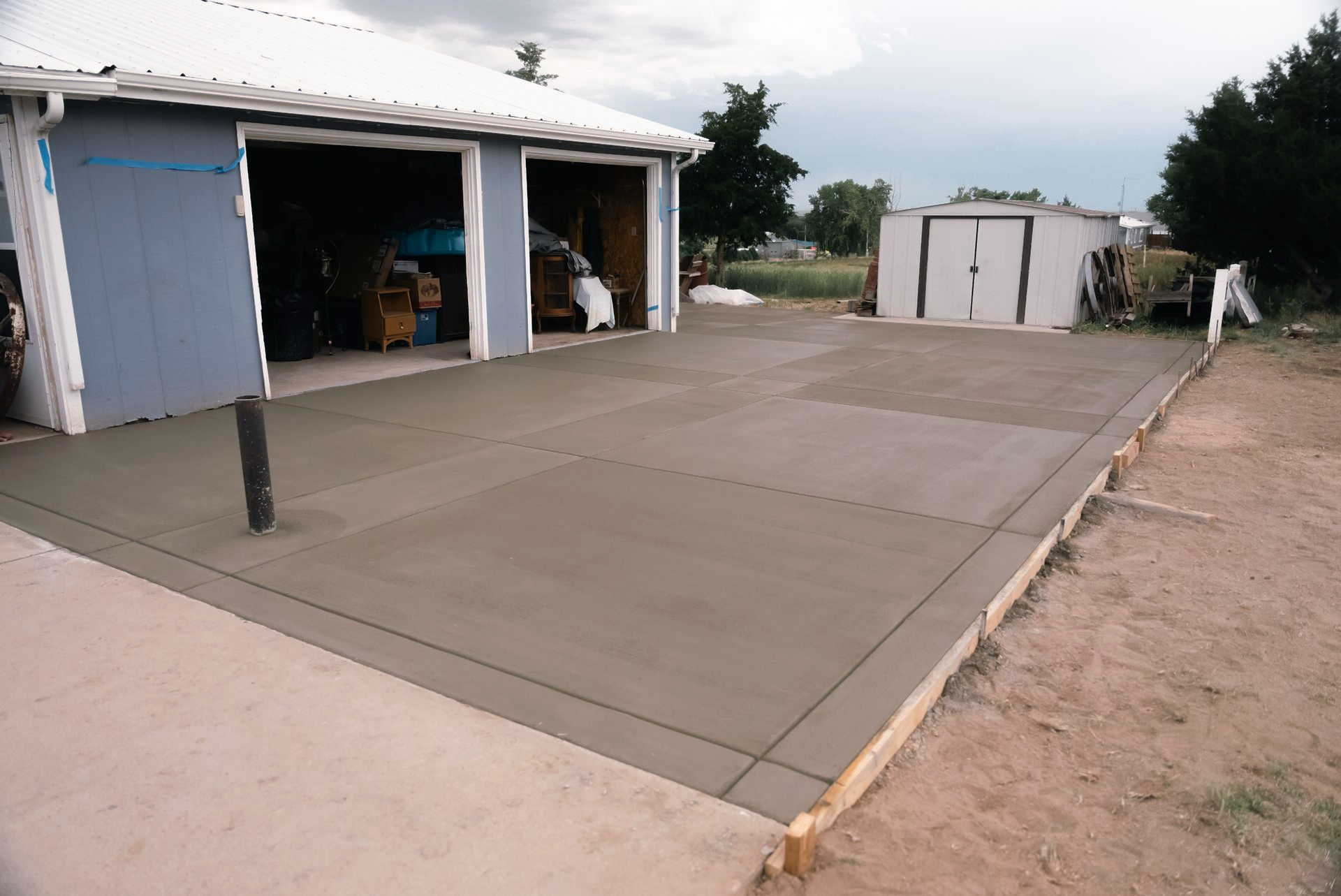 New concrete driveway next to a blue garage and shed on a cloudy day.