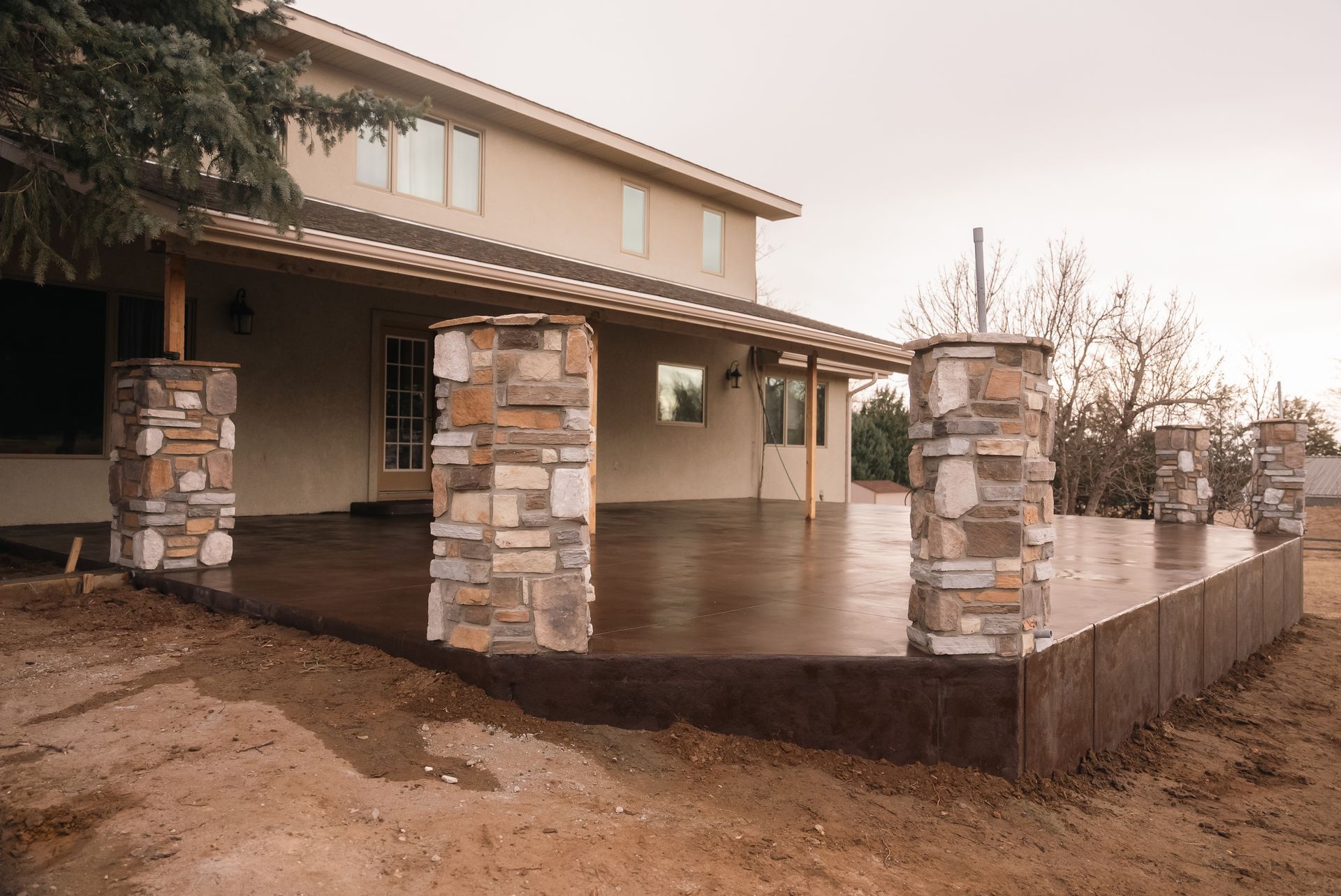 Concrete patio with stone columns and a two-story house in the background. Brown dirt surrounds the patio.