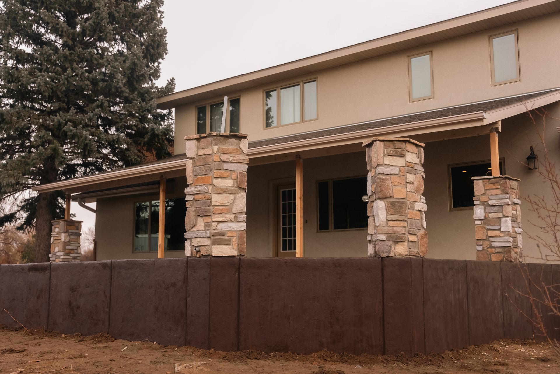 Two-story house with stone columns supporting a covered porch. Brown fence in the foreground, cloudy sky.