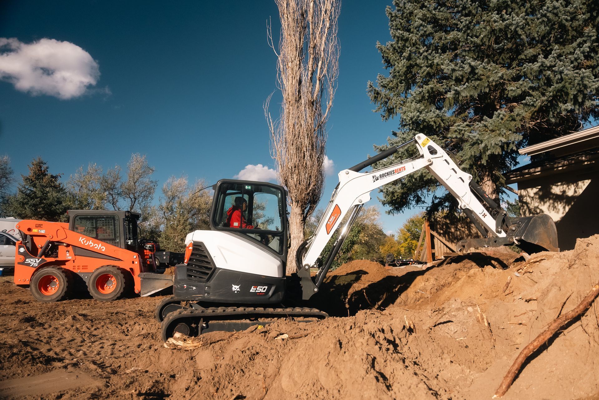 Construction site: Excavator digging in dirt; a skid steer is parked in the background. Bright blue sky.
