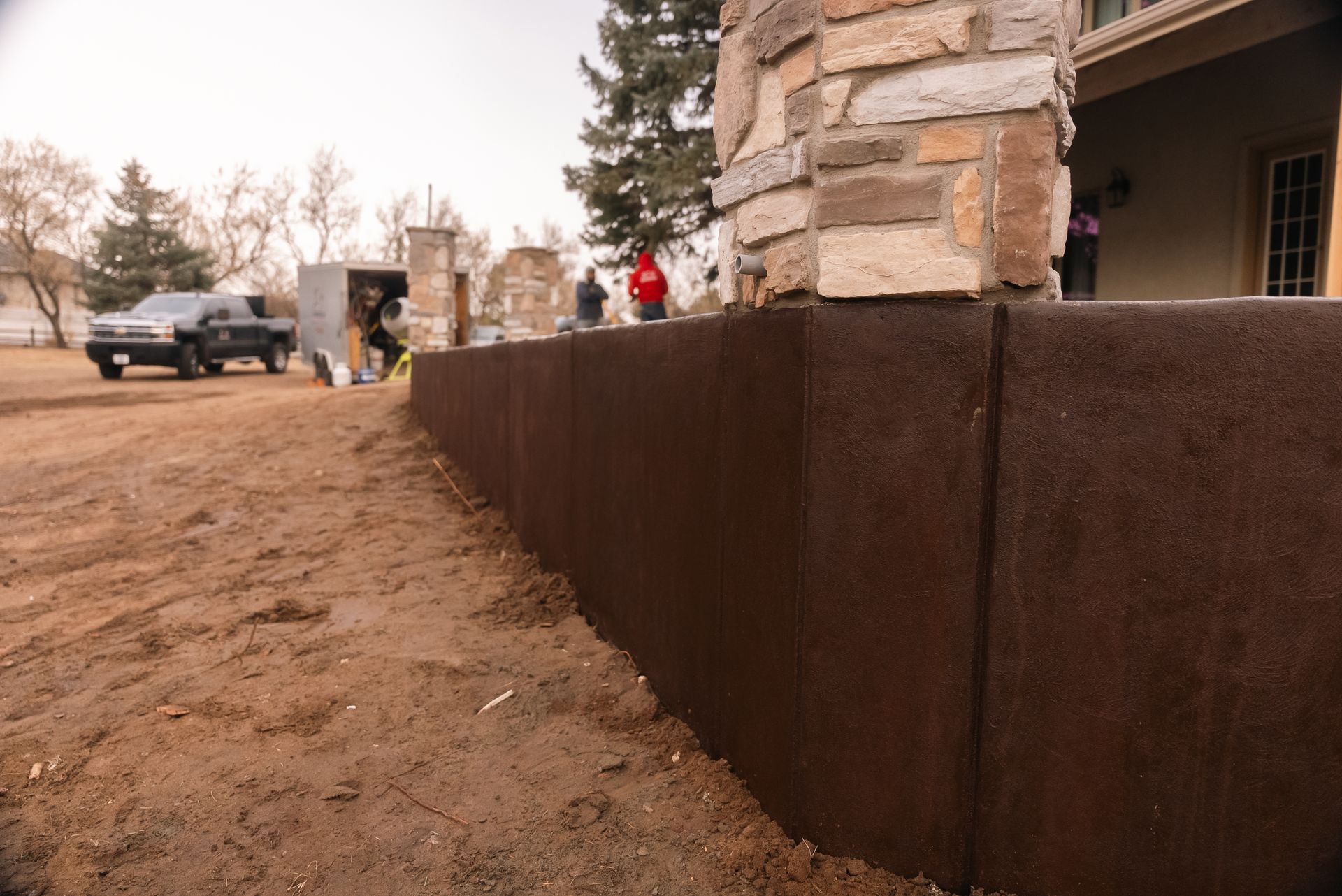 Brown concrete retaining wall in front of a house, with workers and a truck visible in the background.