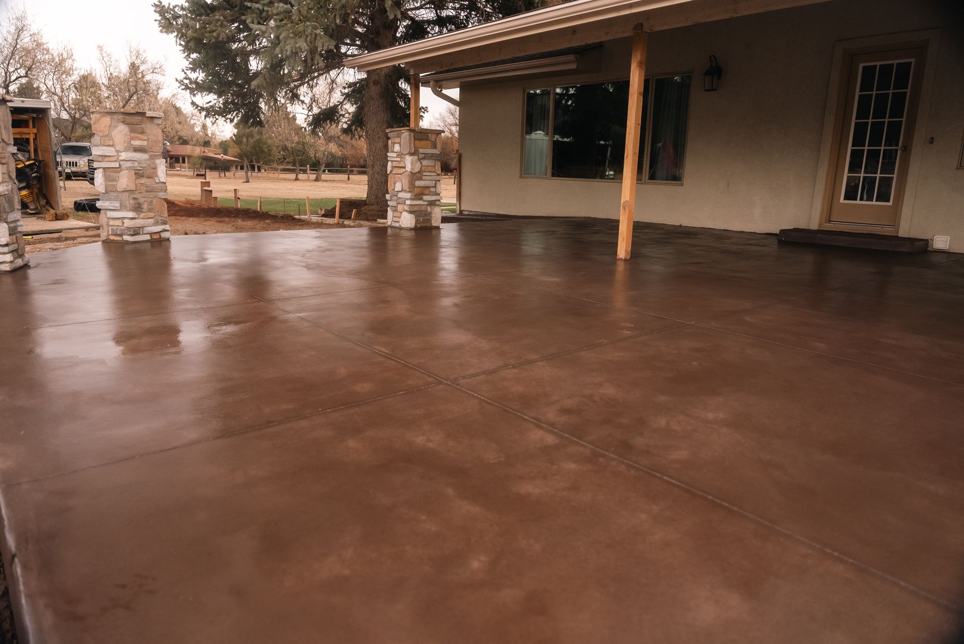 A freshly poured, brown concrete patio in front of a house, with stone pillars and a covered porch.