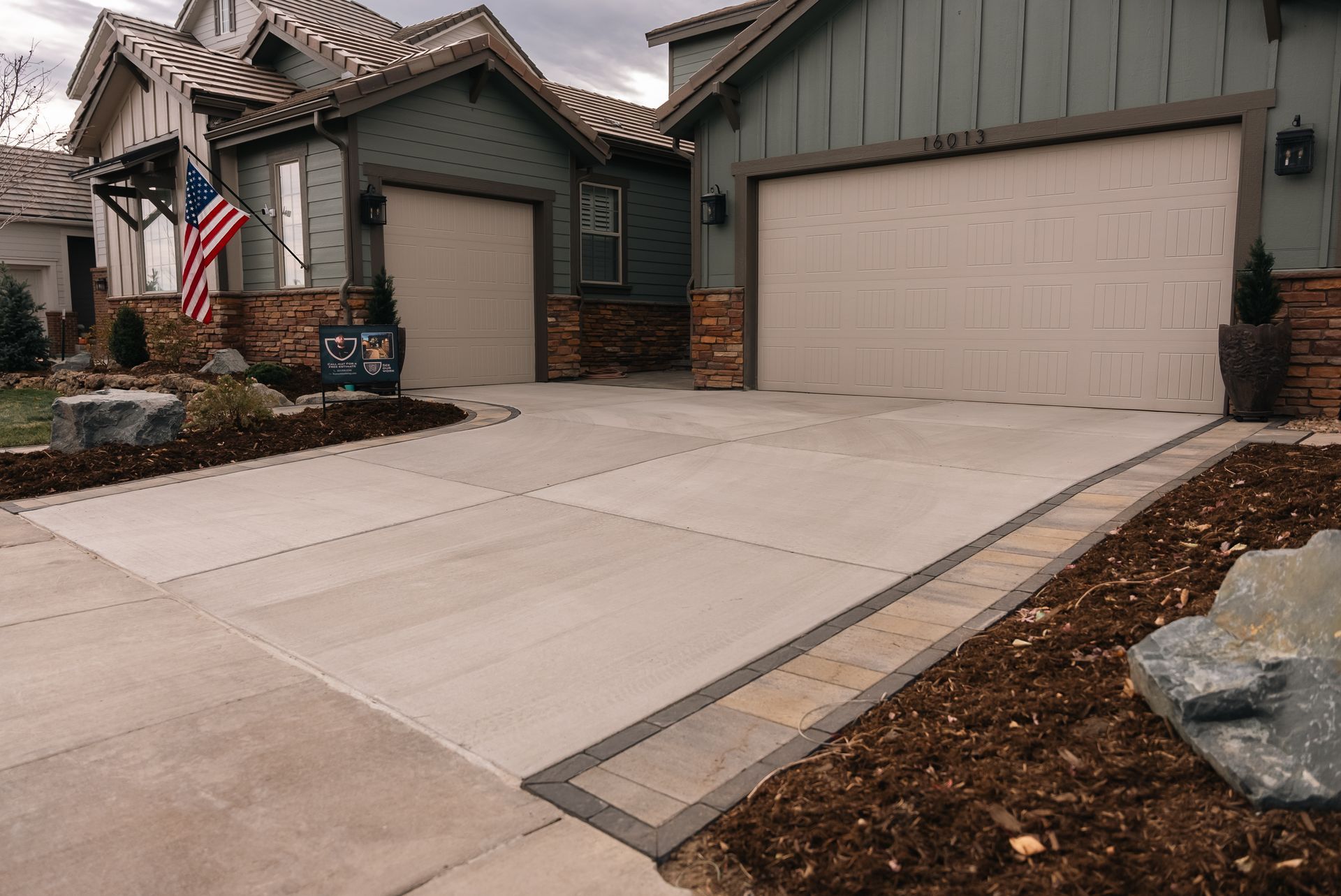 Concrete driveway in front of a house with two garages. The border is made of brick. An American flag is visible.
