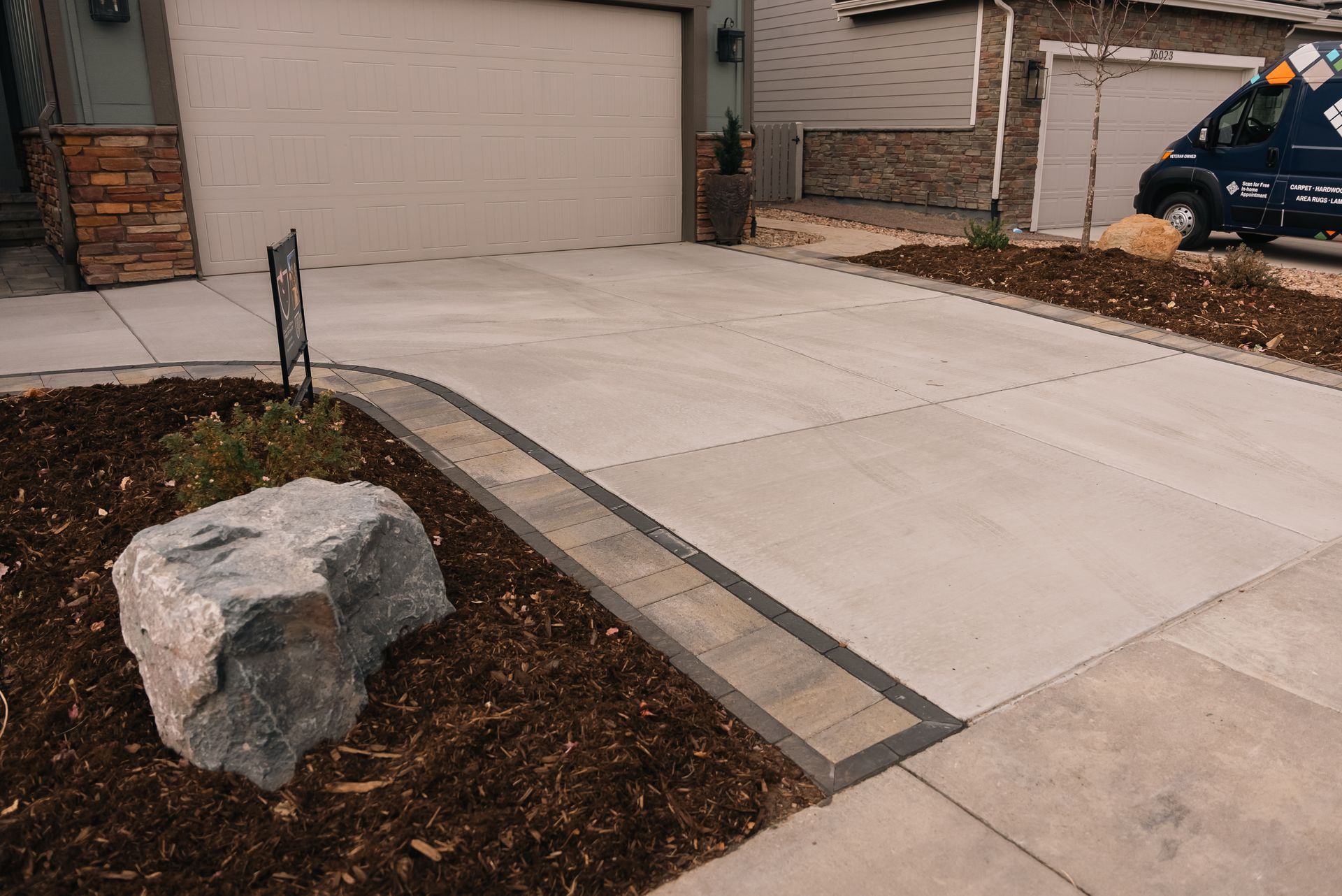 Driveway with concrete slabs, bordered by landscaping and a large rock. Garage and service van are in the background.