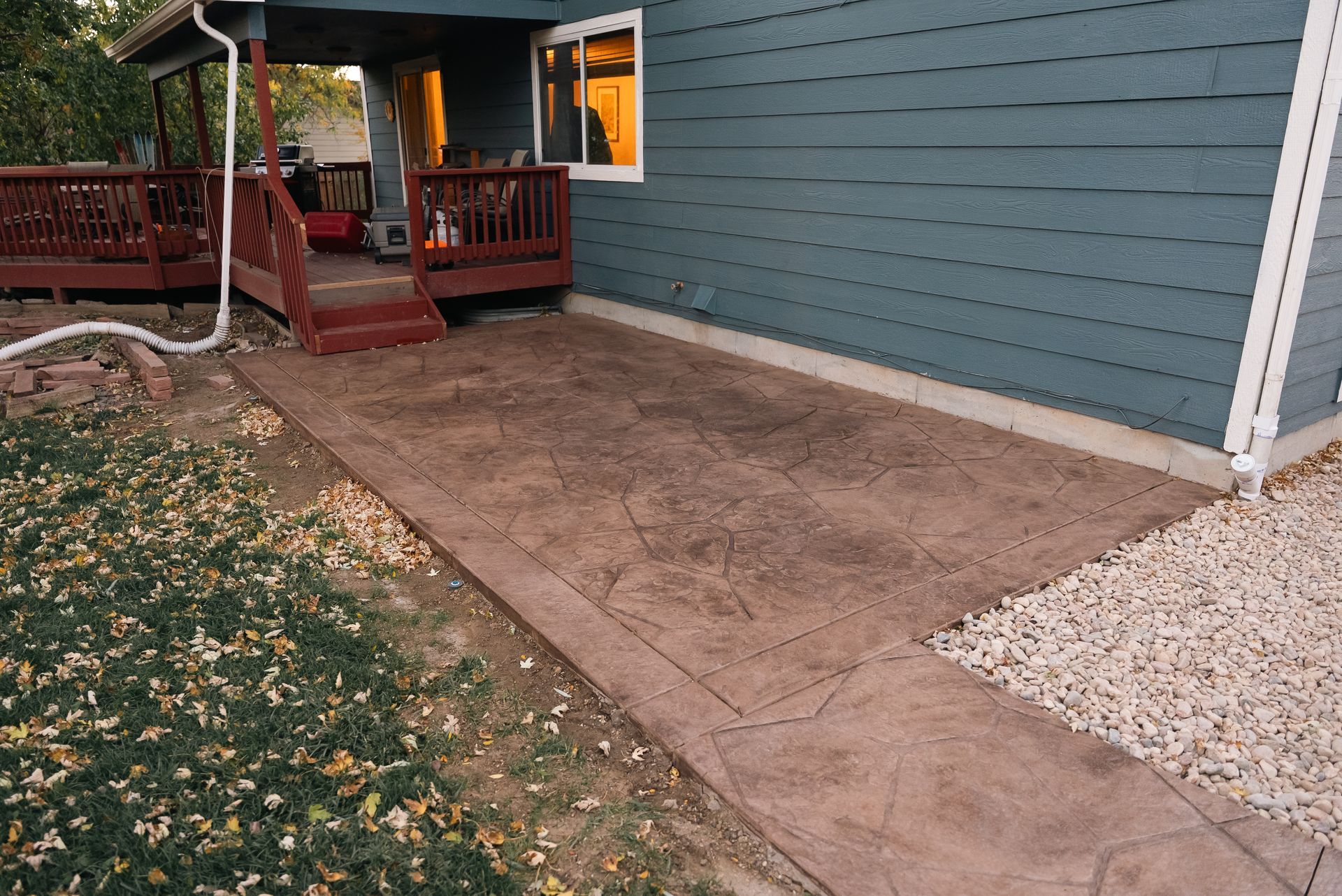 Concrete patio next to a house with a wooden deck and gravel pathway.