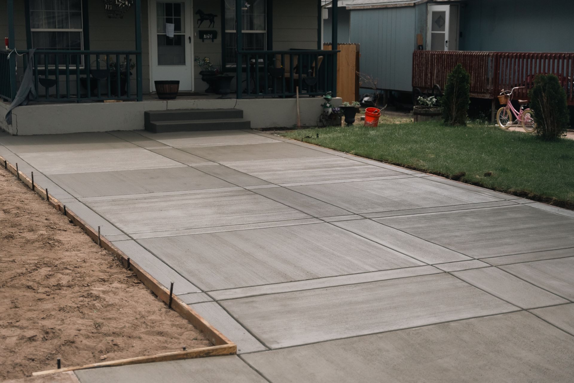 Newly poured concrete driveway with a house in the background. Wooden framing lines the edge, bordering dirt.