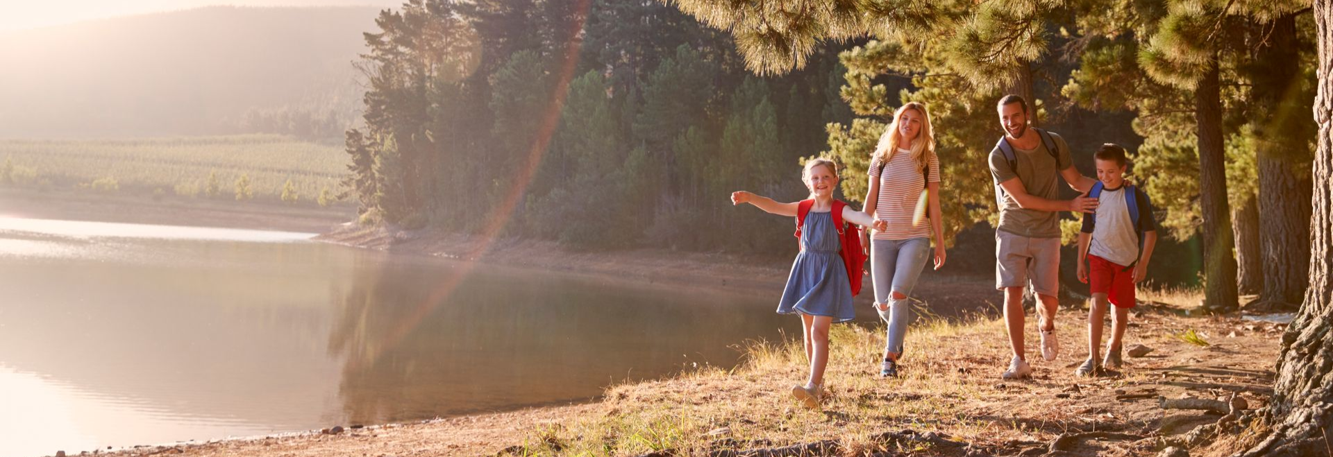 Family walks along a lake, the young girl is running ahead with arms outstretched, trees line the shoreline.
