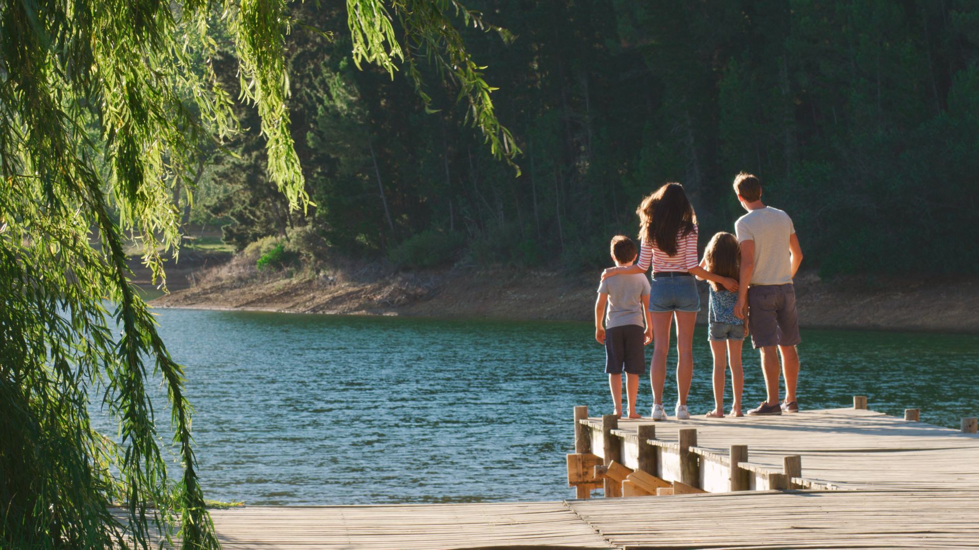 Family standing on a dock, looking out at a lake. Green trees, sunny day.