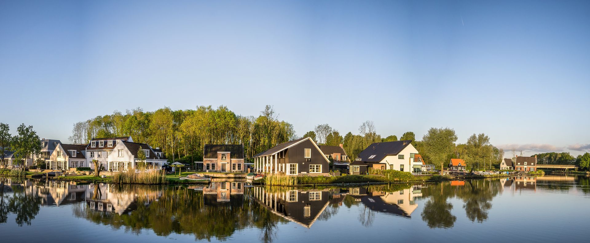 Houses line a lake shore, reflecting in the water. Lush green trees and a clear blue sky complete the idyllic scene.