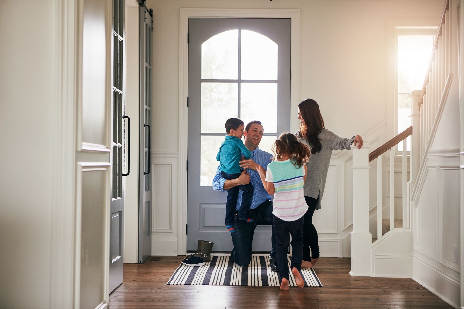 Family in entryway: Father holding a child, two others beside them, near a door and staircase.