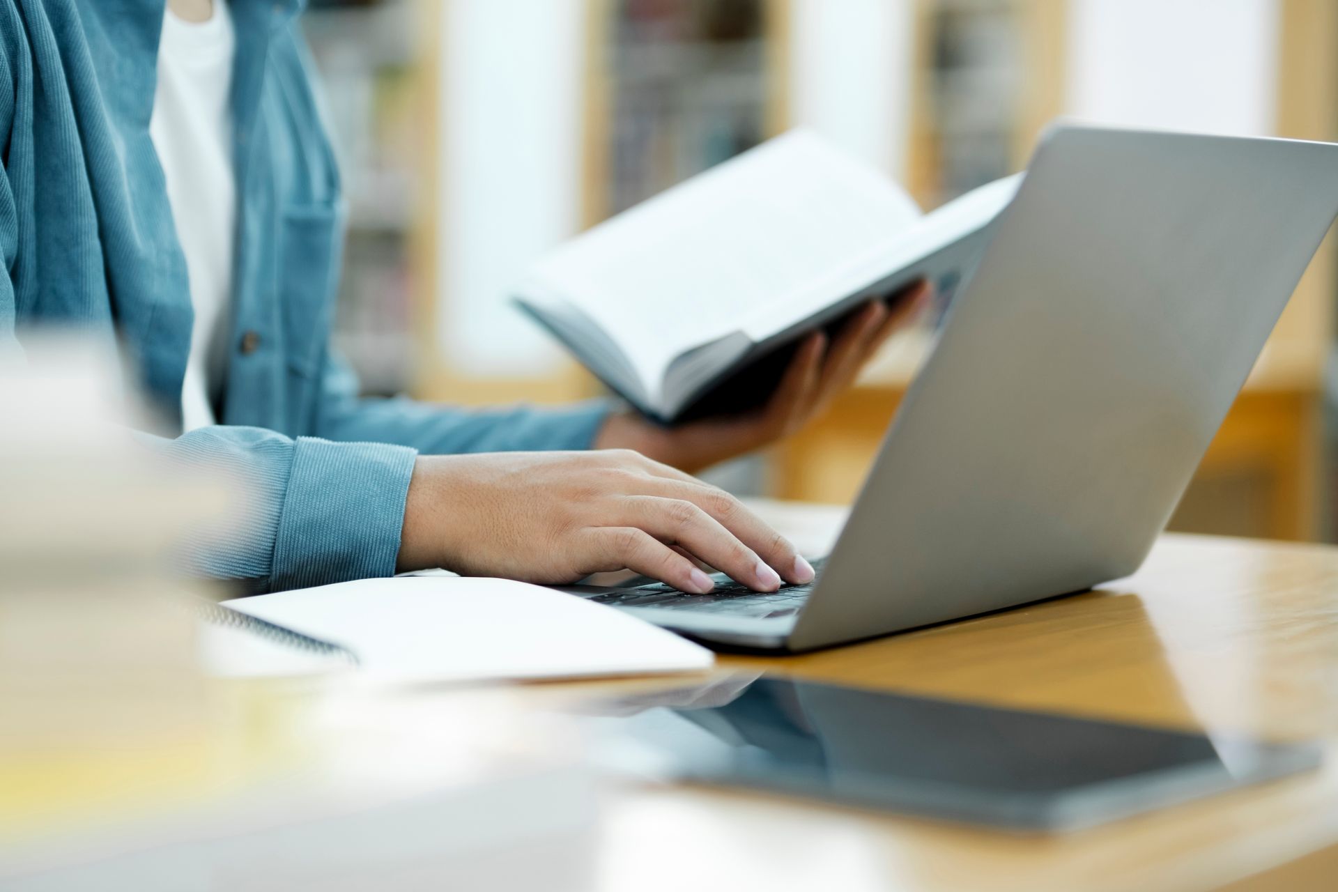 Person at a desk using a laptop and reading a book in a library.