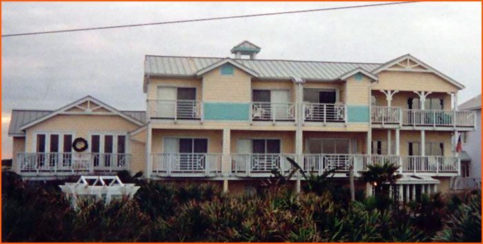 Multi-story yellow house with teal accents, balconies, metal roof, and surrounding foliage.